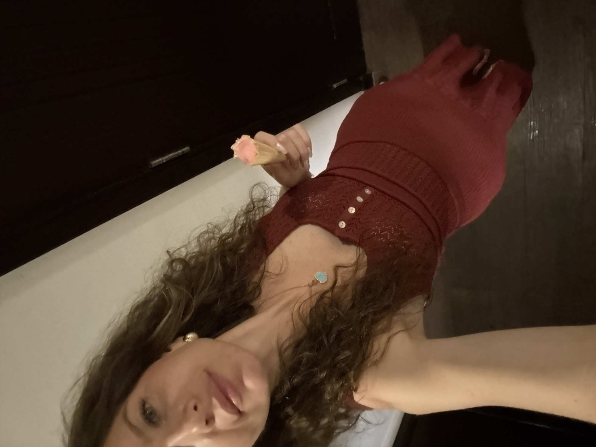 A low-angle, tilted, eye-level indoor photograph shows a woman holding an ice cream cone, situated in the city of Dubai, United Arab Emirates. The woman, with long, dark, curly hair, faces the camera directly with a subtle smile. She wears a deep maroon, ribbed knit dress or top featuring a V-neckline and a line of light-colored buttons down the front bodice. A small, light blue, heart-shaped pendant hangs from a thin gold-colored chain around her neck, and a golden stud earring is visible in her right ear. Her right hand, with light-colored manicured nails, extends towards the viewer, holding a single-scoop, light pink ice cream in a wafer cone. The background consists of a dark, vertical surface on the right, likely a door or wall, next to a narrow vertical strip of white trim or molding on the left. The lighting is ambient and somewhat dim.
