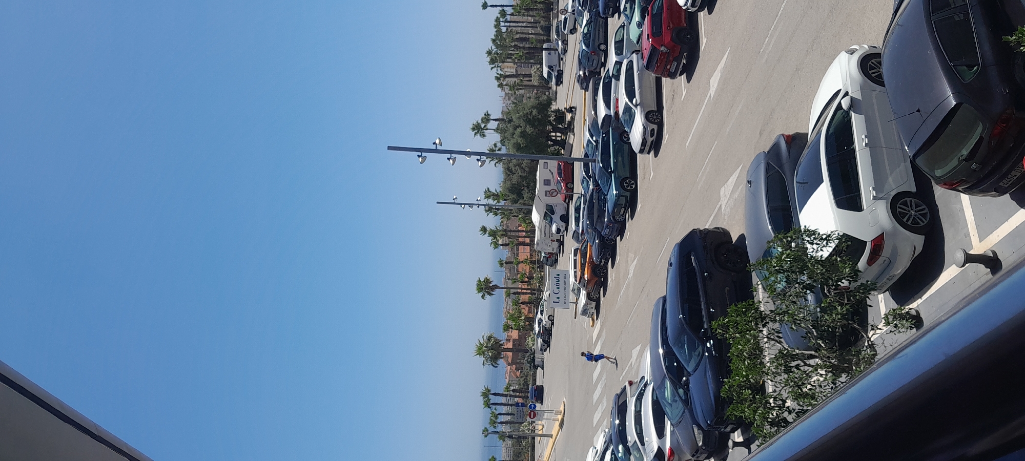 This image captures a wide-angle view of a large, bustling outdoor parking lot under a clear, bright blue sky. The photo appears to be taken from an elevated position, possibly a balcony or higher floor, looking down and across the parking area.
Numerous cars of various models and colors, including white, black, grey, red, blue, and orange vehicles, as well as a few camper vans, are parked in neatly delineated spaces. The parking lot pavement is asphalt with white painted lines for parking bays and a crosswalk. Strong shadows cast by the vehicles indicate bright sunshine, suggesting midday or early afternoon.
In the middle ground, a lone individual dressed in blue is walking across a crosswalk. Several tall, modern streetlights with multiple fixtures illuminate the parking area. Beyond the parking lot, rows of lush palm trees line the area, interspersed with some green foliage. Behind the trees, residential or commercial buildings with light-colored roofs are visible, and in the distance, a hazy blue expanse of what appears to be the sea or a large body of water can be seen.
A prominent white sign with dark lettering stands in the parking lot, clearly displaying "LA CAÑADA" with "GALERÍAS COMERCIALES" written underneath. This text confirms the location as La Cañada shopping center in Marbella, Spain. The foreground of the image shows a dark railing or architectural element from the vantage point. The overall scene depicts a sunny day at a busy commercial center in a Mediterranean coastal region.