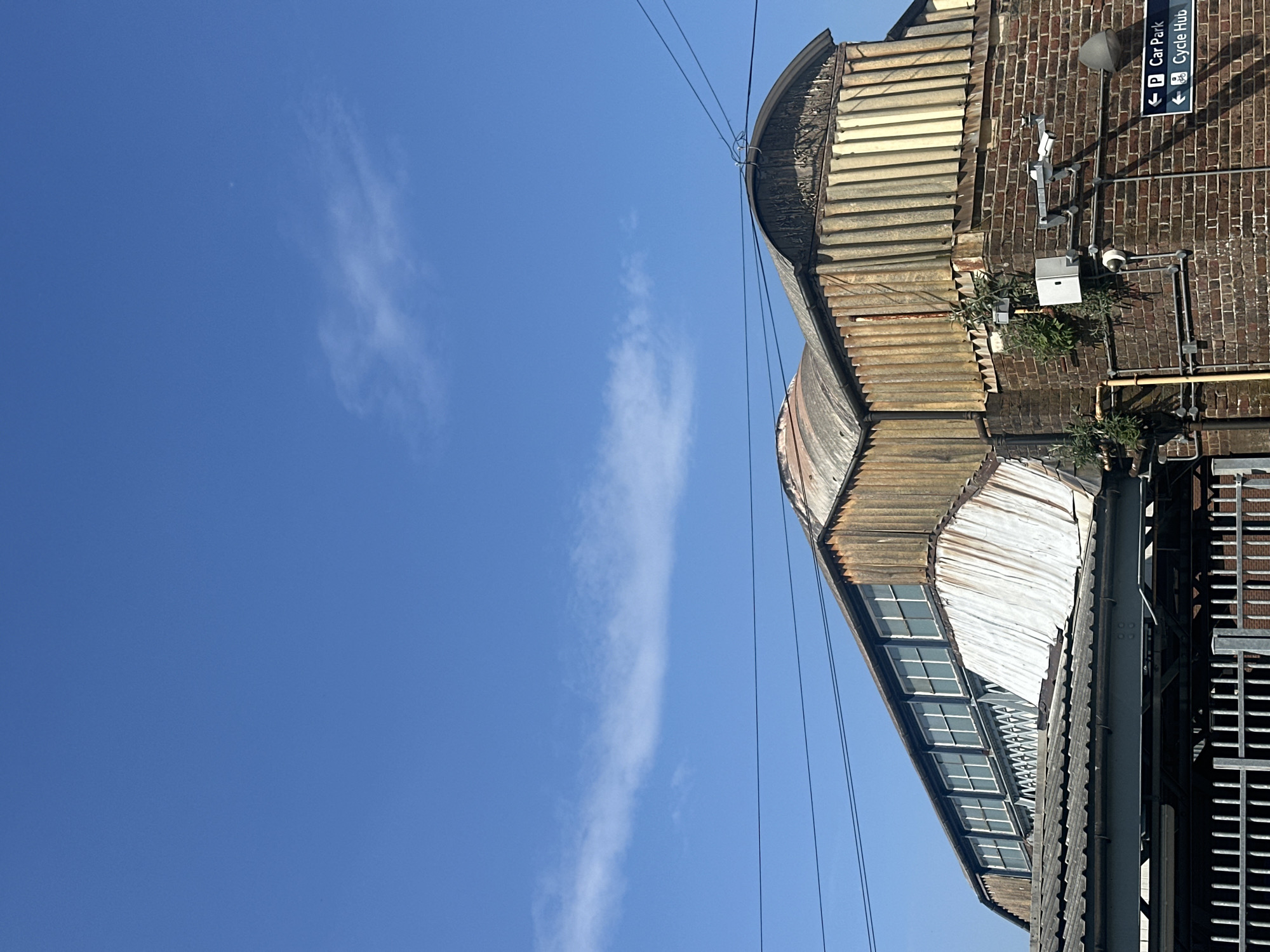The image, when viewed upright, displays an architectural detail of a building under a bright blue sky. The left side of the frame is dominated by a clear, vibrant blue sky, featuring a few wispy, elongated white clouds. Several thin, dark wires stretch horizontally across the upper portion of the sky, connected to a structure on the right.
The right side of the photograph features a weathered industrial-style building. Its roof is characterized by a series of curved, corrugated metal sheets, showing signs of age with various tones of grey, brown, and hints of rust. Below this, another section of the building reveals a façade with multiple small-paned windows, suggesting a clerestory or sawtooth roof design that allows light into the interior.
A reddish-brown brick wall forms a prominent part of the building's exterior. Mounted on this wall are several fixtures: two security cameras, a grey utility conduit, a white electrical box, and a grey speaker or light. Small patches of green vegetation are growing directly from the brickwork in various spots.
A notable detail on the brick wall is a blue directional sign with white text and symbols. It reads "P Car Park" with an arrow pointing left, and "Cycle Hub" with an arrow pointing diagonally down-left. Below this, a dark, grid-like metal structure is visible in the lower right, possibly part of a walkway, bridge, or station platform.
The scene is well-lit by direct sunlight, casting clear shadows and indicating it is daytime. There are no people visible, and no specific activity is taking place. The presence of "Car Park" and "Cycle Hub" signs, along with the industrial architecture and the given location context of "Waverley, United Kingdom," strongly suggests this is part of or adjacent to Edinburgh Waverley railway station.
