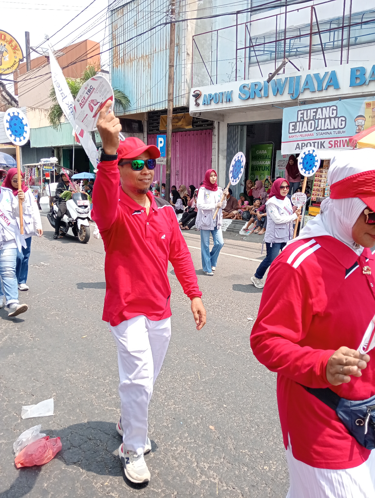 In Demak, Indonesia, a parade is underway on a street lined with buildings, including the "APOTIK SRIWIJAYA BA" pharmacy. Participants, many wearing red and white, carry signs featuring a depiction of the coronavirus. The central figure, a man in a red shirt and white pants, raises a sign and wears sunglasses. Other individuals in the procession are also visible, some holding similar signs and wearing hijabs. A motorcycle is present in the background, and litter is scattered on the asphalt road. The sky is clear, indicating daytime.