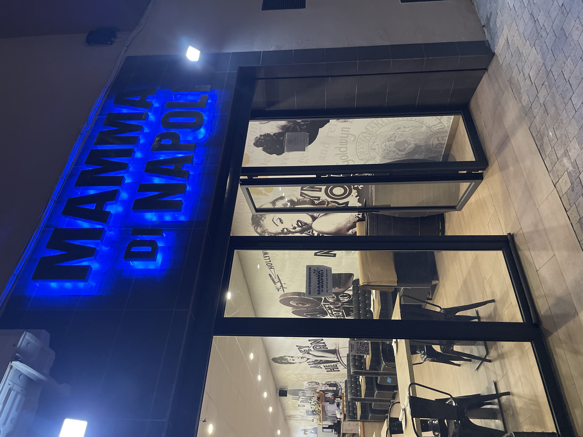 The image shows the exterior of a restaurant called "MAMMA DI NAPOLI" in La Línea De La Concepción, Spain. The restaurant has a prominent blue neon sign above the entrance. Through the glass doors and windows, the interior is visible, revealing dining tables and chairs, and murals on the walls depicting classic Hollywood imagery such as Audrey Hepburn and Marilyn Monroe. A QR code is visible on a sign inside the window. The lighting suggests it is nighttime.