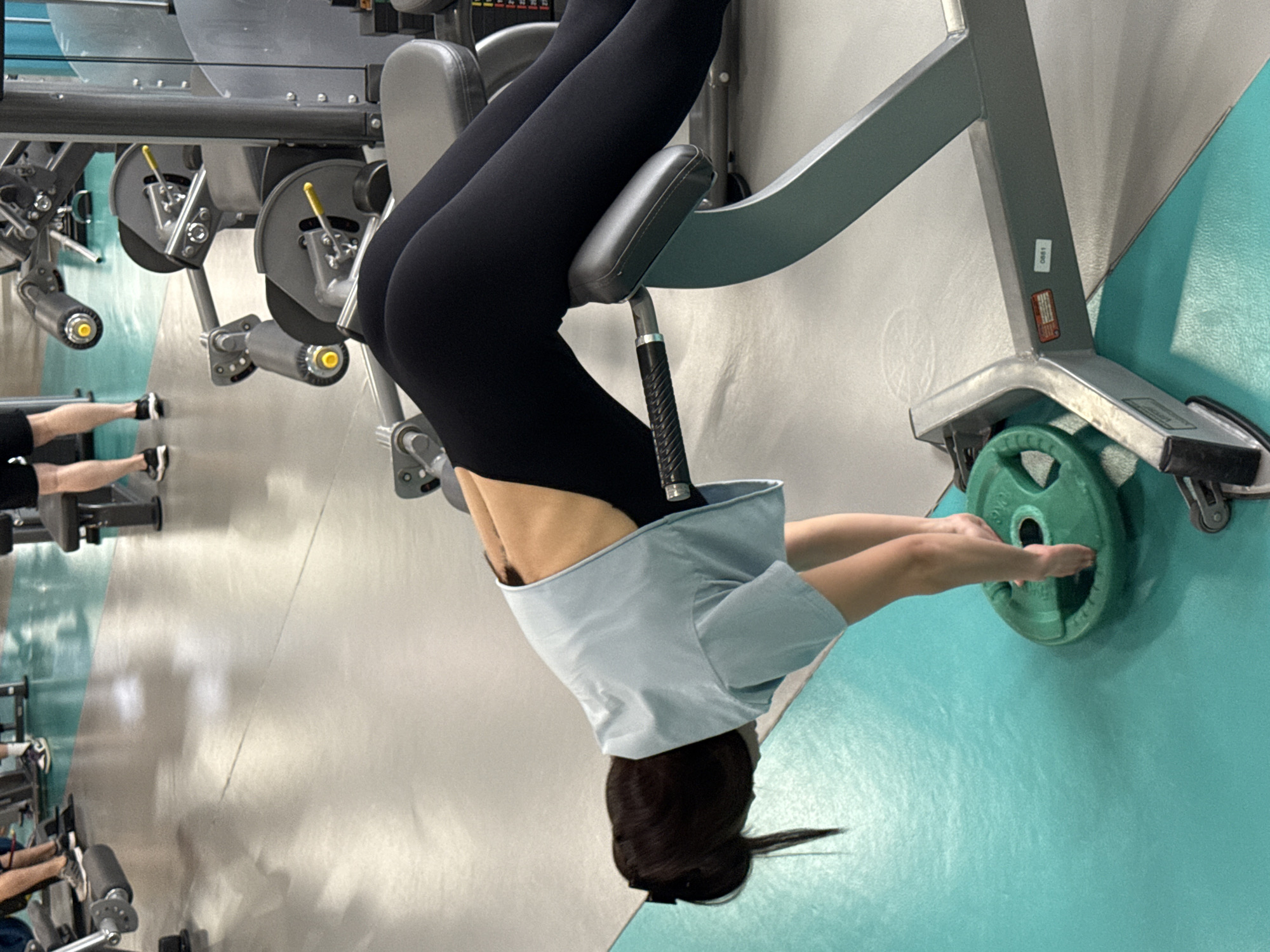 A woman is using a workout machine in a gym. The woman is wearing black leggings and a light blue t-shirt. Her legs are suspended in the machine, and her hands are holding onto a green weight plate. The gym has a light gray floor and turquoise blue walls. Other gym equipment is visible in the background. This scene is occurring inside a gym located in an unknown part of Yekaterinburg, Russia.
