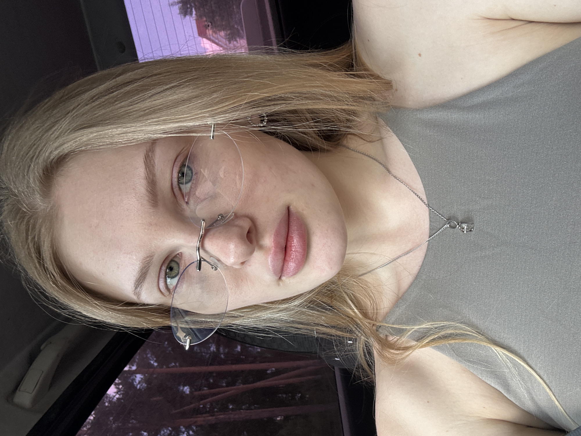A young woman with blonde hair and wearing glasses is shown in a selfie. She is wearing a gray top and a silver necklace with a small pendant. The background includes a car window and what appears to be trees. There is no specific information that confirms the location is Perm, Russia.