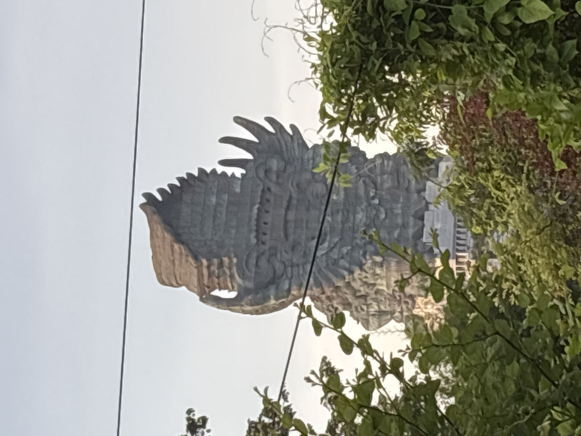 The image displays a colossal statue or sculpture, likely depicting the head and upper torso of a mythical bird or deity, set against a light, overcast sky. The sculpture is predominantly grey with some brown accents, intricately textured with carved details resembling stone or metal. Visible features include a prominent beak-like mouth, ornate crown-like elements or elaborate plumage on the head and neck, and a patterned, block-like surface.
Dense green foliage, including leaves, branches, and vines, frames the sculpture, primarily along the right side of the frame, partially obscuring some of its features. Thin, dark lines, possibly power lines or wires, traverse vertically across the left side of the image, cutting across the sky and a small portion of the sculpture.
The background consists of a uniform, light grey sky, suggesting an overcast day or diffused lighting. The scene captures a static monumental work of art, with no specific activity taking place. The immense scale and detailed craftsmanship of the sculpture strongly indicate a significant cultural or tourist landmark, consistent with the Garuda Wisnu Kencana Cultural Park located in Kuta Selatan, Indonesia. No text is visible in the image.