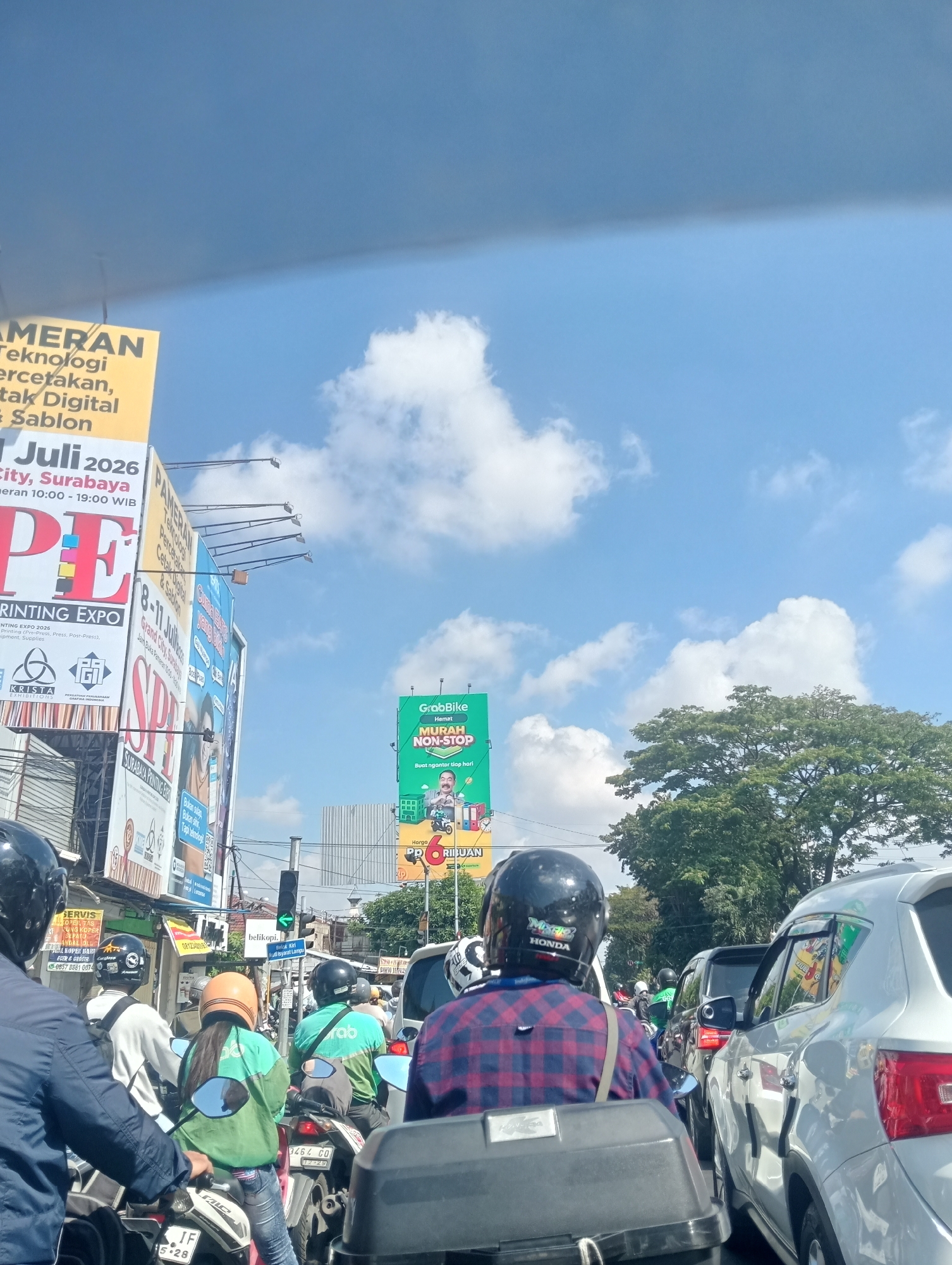 The image shows a congested street scene in Surabaya, Indonesia. Several motorcycles and cars are stopped in traffic. Many people are wearing helmets, suggesting they are riding motorcycles. There are several large billboards visible along the roadside. One billboard is for a "Printing Expo" with dates July 1-8, 2026, and mentions "Grand City, Surabaya". Another billboard advertises "GrabBike" with the slogan "MURAH NON-STOP". The sky is blue with some white clouds. The overall impression is of a busy urban environment on a sunny day.