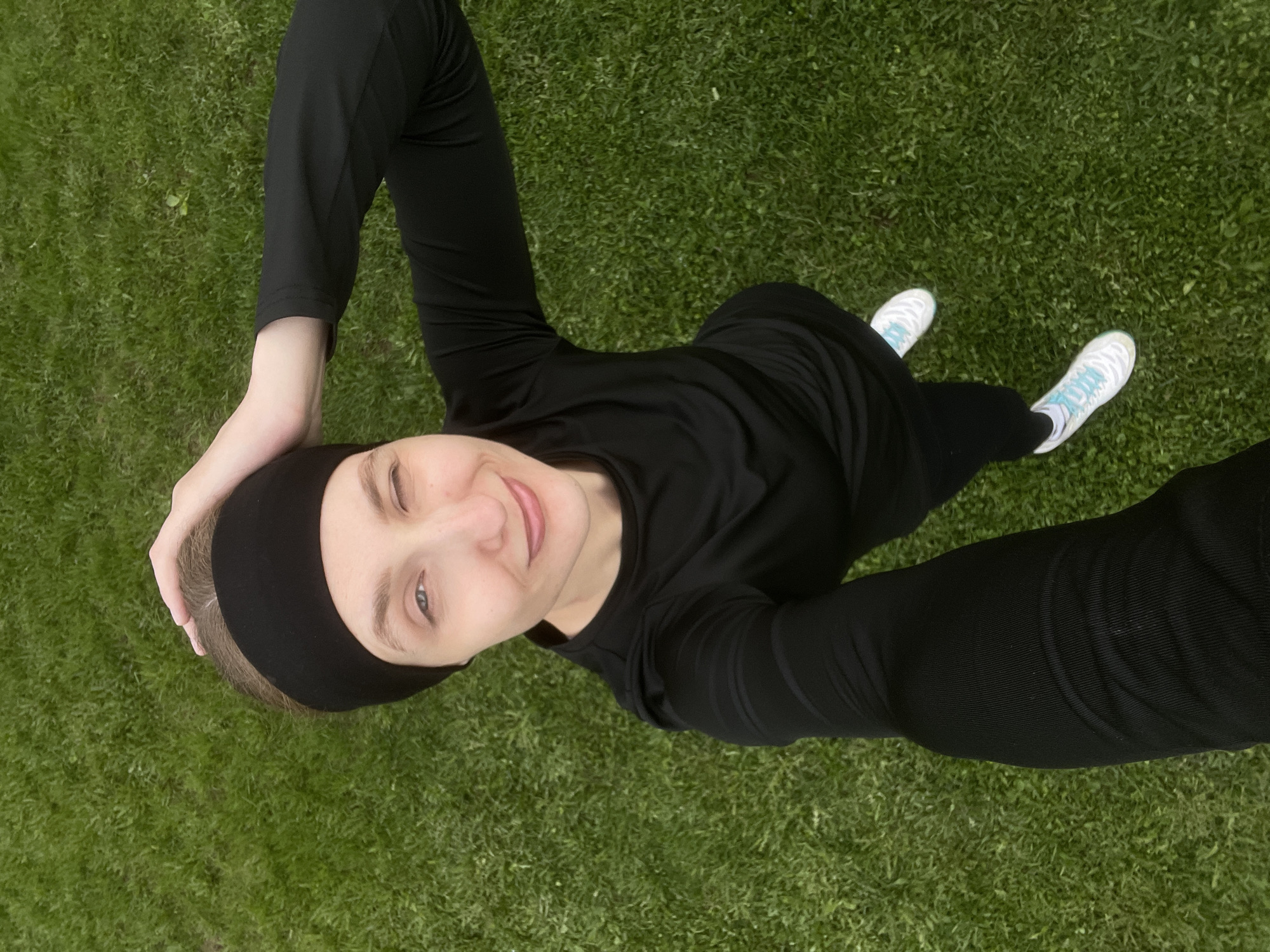 A woman is standing on a lawn of green grass, taking a selfie from a high angle. She is wearing a black long-sleeved top, black pants, and white sneakers with blue details. She has a black headband on her head and is smiling, winking one eye. Her right hand is on her head, and her left arm is extended, holding the device taking the picture. The location is unknown.