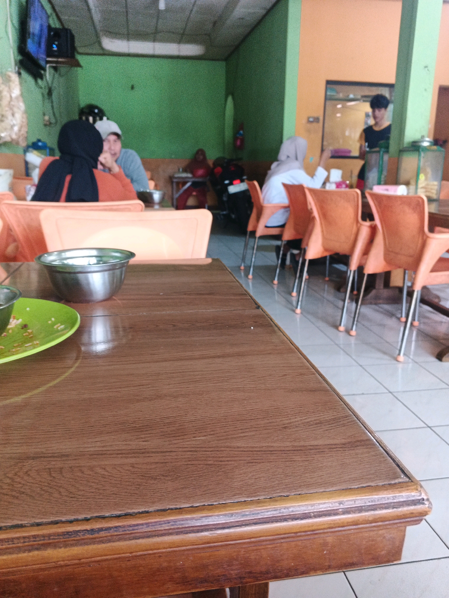 The image depicts the interior of a simple restaurant in Ungaran, Indonesia. A wooden table occupies the foreground, with a green plate and a metal bowl visible on its surface. In the background, several people are seated at tables, some wearing headscarves, suggesting a predominantly Muslim environment. The walls are painted in green and orange. To the right, a server stands with a tray. The overall scene suggests a casual dining establishment.
