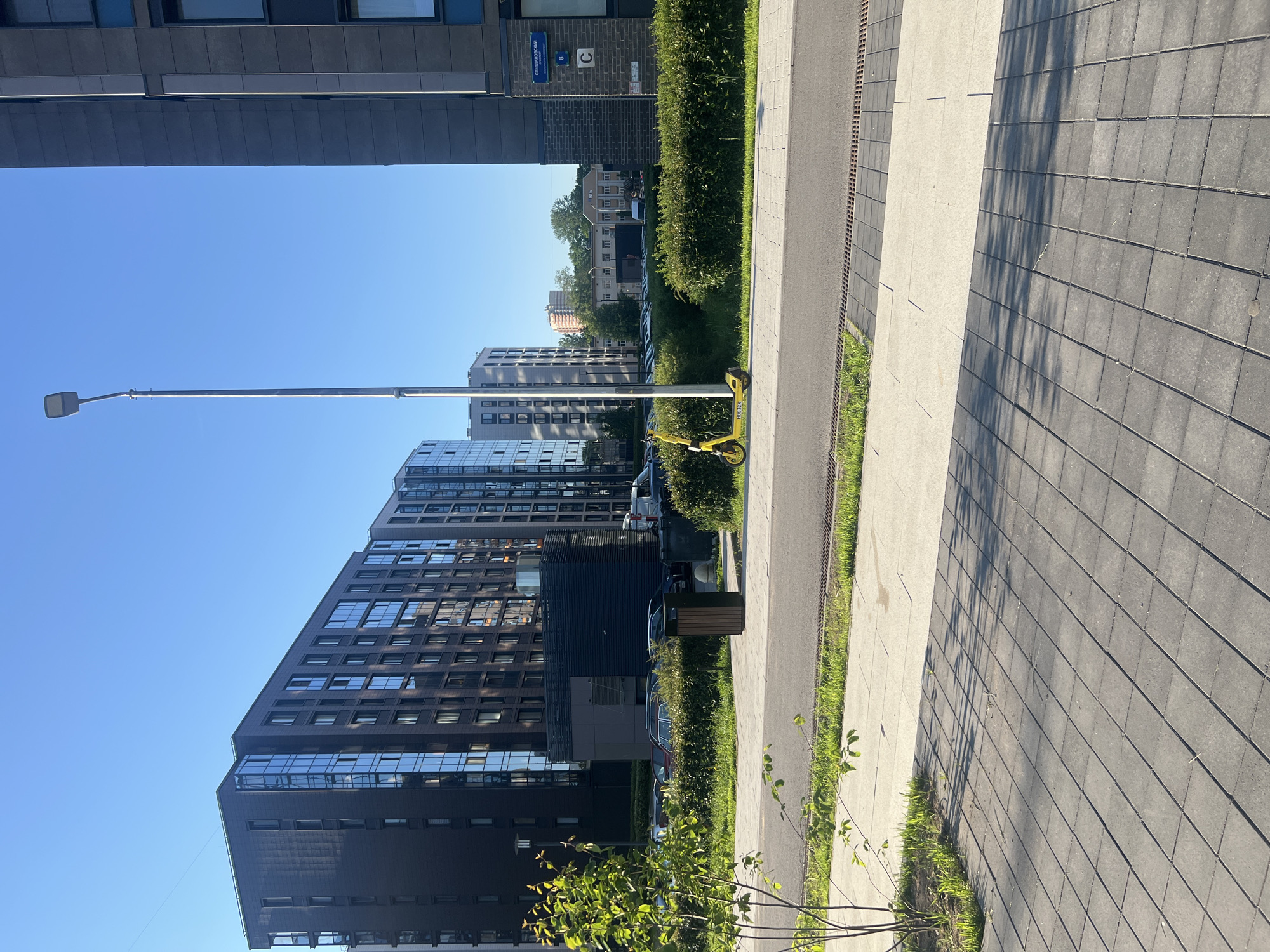 The image depicts a bright, sunny urban street scene. The view is along a wide, paved pedestrian walkway composed of contrasting light and dark grey square paving stones, with strong shadows cast across it. To the left of the walkway, several modern, multi-story buildings with dark facades and numerous reflective windows rise towards a clear blue sky. One of these buildings has a distinct angular or stepped architectural design.
Separating the walkway from a parking area and a narrow road beyond, is a well-maintained, lush green hedge. Behind this hedge, several parked cars are visible, and further in the distance, more buildings can be seen, including a lighter-colored one with "ВТБ" prominently displayed on its roof. A tall, slender grey street lamp stands near the hedge. Parked conspicuously near the lamp post and hedge is a yellow electric kick-scooter, with the word "KICKSHARING" visible on its side. A dark brown trash can is also placed alongside the hedge. Grass and small green plants grow along the edges of the walkway and in the foreground.
No people are visible, suggesting a quiet moment. The clear blue sky and strong shadows indicate a bright, sunny day, likely during the morning or afternoon. Visible text on a blue street sign attached to a building reads "СВЕТЛАНОВСКИЙ" (Svetlanovsky) and "8 С", providing a specific location cue consistent with a Russian urban environment. The "ВТБ" logo on the distant building further supports this context.
