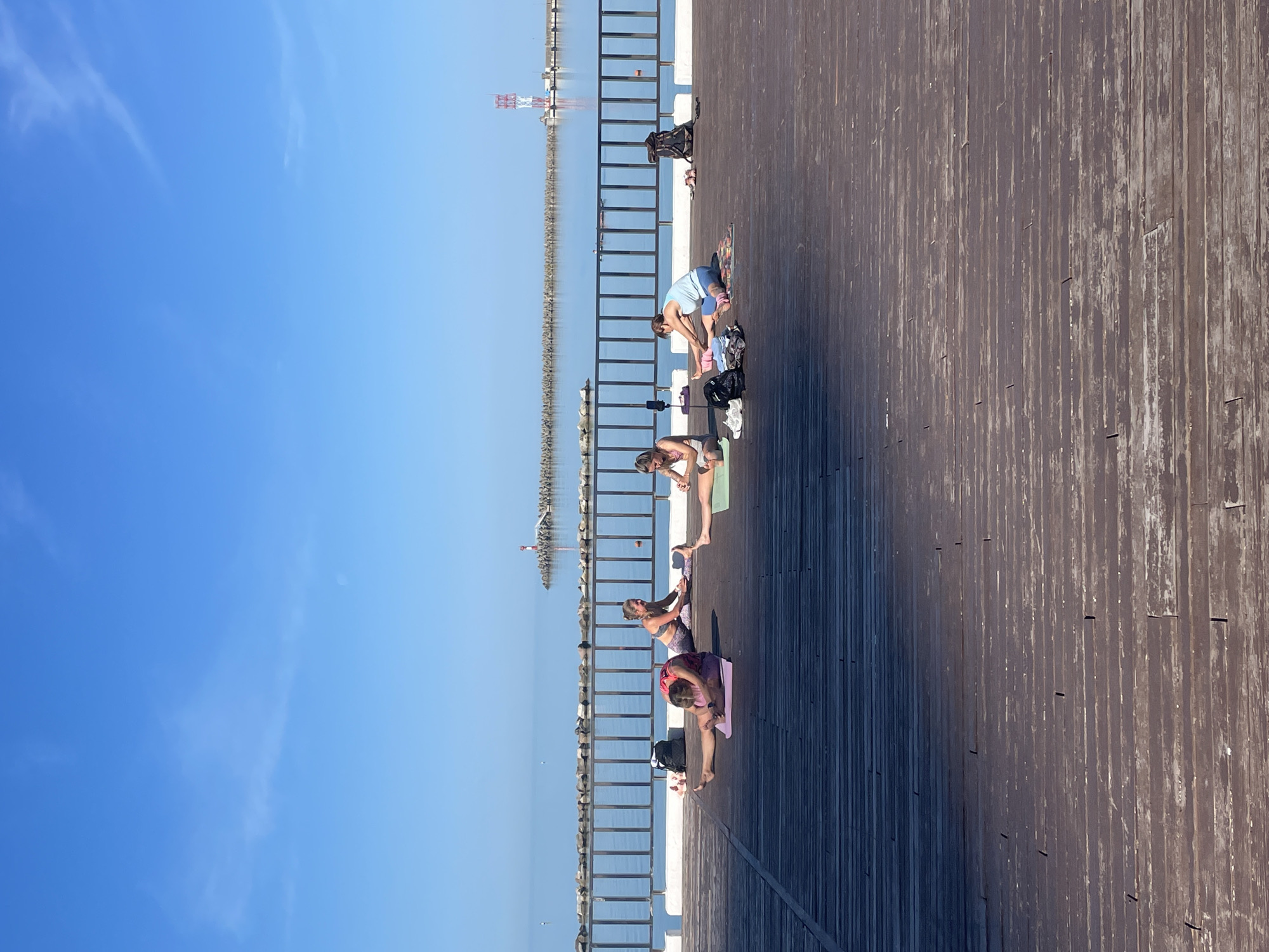 The image depicts a serene outdoor yoga session taking place on a wide wooden pier in Sochi, Russia. Four women are positioned along the pier, each on a yoga mat, engaged in various stretching and bending poses. They are dressed in activewear, and personal items like water bottles and small bags are placed near their mats.
The pier is constructed of dark, weathered wooden planks and extends into the right side of the frame. A black metal railing runs along the edge of the pier, separating it from the calm, blue sea. To the left of the railing, the sea stretches to the horizon, where a long, low breakwater or jetty is visible. Several structures, possibly navigational markers or small lighthouses, can be seen along this distant barrier, including one with distinctive red and white stripes.
Above the horizon, the sky is a clear, vibrant blue with only a few faint, wispy clouds. The bright sunshine indicates a clear day, likely late morning or early afternoon, casting distinct shadows from the railing and the practicing individuals onto the wooden surface of the pier. The scene conveys a sense of peaceful activity and connection with nature. No discernible text is visible in the image.