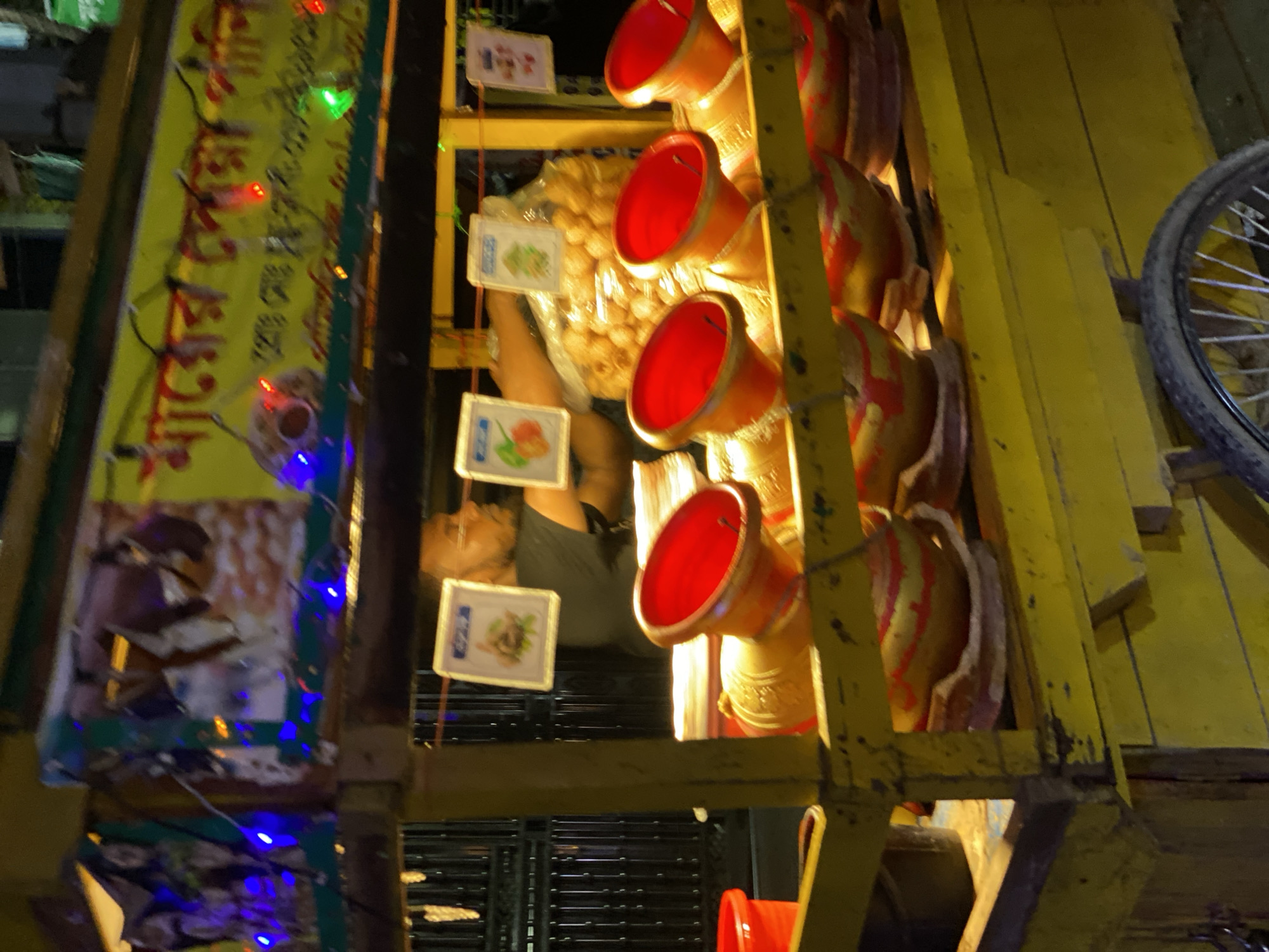 This image captures a vibrant street food stall at night, likely in Dhaka, Bangladesh, given the visible Bengali text. The scene is illuminated by numerous artificial lights, including colorful fairy lights (red, green, and blue) strung along a large yellow sign, and brighter internal lighting that highlights the food display.
At the center of the stall, a person, presumably the vendor, is visible lying down or resting behind the counter, wearing a dark top. Their arm is prominently seen. The primary subject of the stall appears to be a popular South Asian snack resembling *pani puri* or *golgappe*. A large clear bag or container holds a multitude of small, round, golden-brown fried shells, which are the main component of these dishes.
The stall features a distinctive display of serving containers. Several red, trumpet-shaped containers are arranged vertically, likely used for holding different flavored waters (*pani*) that are poured into the puri shells. To the right, brown or tan, possibly clay or ceramic, bowl-like containers are also stacked, perhaps for serving the filled puri. The entire food display is housed within a sturdy yellow wooden or metal framework.
On the left side, a prominent yellow sign adorned with red Bengali script is visible. Although much of the text is obscured or out of focus, some characters resembling "চটপটি" (Chotpoti, a popular street snack) and "খাবার" (Khabar, meaning food) can be partially deciphered, consistent with the food items on display. Smaller, square white signs featuring images of various food items, possibly menu options or ingredient pictures (such as something green, and a red/orange item with a green stem), hang from strings, adding to the visual appeal of the stall.
In the far right of the frame, part of a bicycle wheel is visible, suggesting the stall might be a mobile cart or located near parked bicycles. The overall atmosphere is that of a bustling urban night market or street, typical of many South Asian cities.