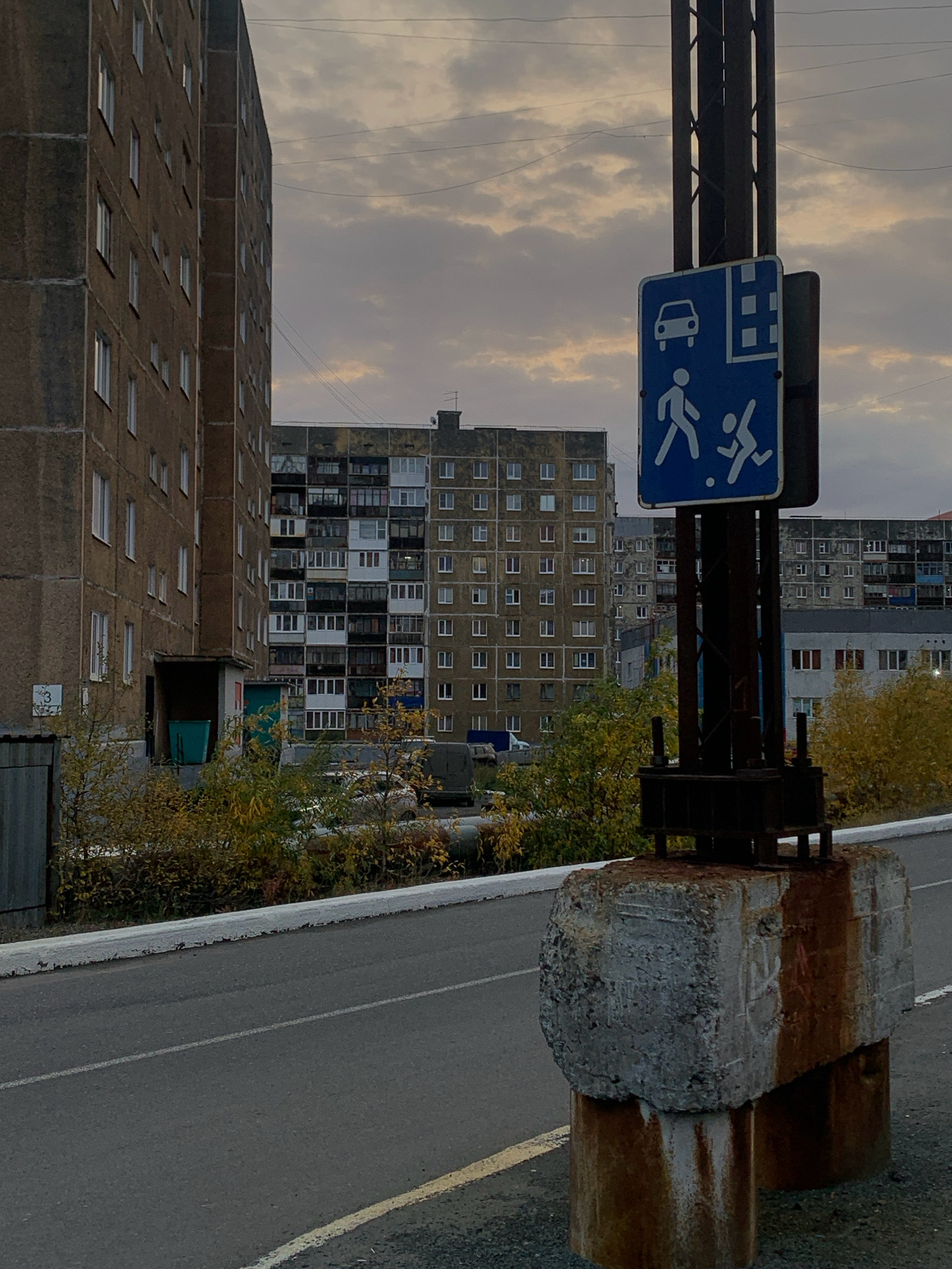The image depicts a street scene in Norilsk, Russia, characterized by austere, Soviet-era apartment buildings under a cloudy, overcast sky. In the foreground, a dark asphalt road with a yellow line marking curves away. To the right, a concrete foundation stained with rust supports a metal pole with a blue traffic sign. The sign displays a white car symbol, a pedestrian figure, and a child in motion. Further down the road, rows of uniform, multi-story apartment buildings dominate the landscape, their facades weathered and utilitarian. Autumn foliage in shades of yellow and brown adds a touch of color to the otherwise monochromatic environment. Several parked vehicles are visible in the background, along with power lines crisscrossing the sky. The overall impression is one of a stark, industrial city in a northern climate.