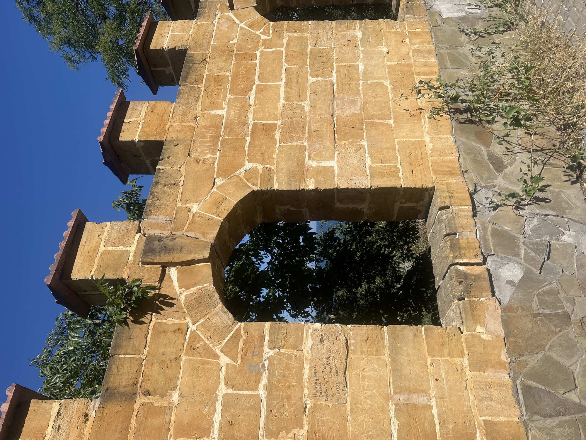 The image shows a section of an old stone wall, likely part of a historical fortification or building, in Odesa, Ukraine. The wall is constructed from large, roughly hewn, yellowish-brown stone blocks. The top of the wall is crenellated with regularly spaced square turrets capped with red terracotta tiles. In places, greenery, such as vines and small trees, is growing on and over the wall.
There are at least two arched openings in the wall, suggesting it may have served as a defensive structure or an entranceway. Through one of the arches, dense green foliage and a glimpse of a blue sky are visible, hinting at the natural surroundings outside the wall, possibly overlooking the sea or a park.
The surface of the stone blocks is covered with numerous inscriptions and carvings, suggesting it has been a site of public access or historical significance where people have left their marks over time. Some of these carvings are clearly legible, with dates like "2026" and names or place names like "Odessa" and "Hacibey."
The lower part of the wall transitions to a base constructed from smaller, irregular, grey and brown stones, creating a textured foundation. Dry grass and weeds grow at the base of the wall, indicating an outdoor environment. The sky above is a clear, vibrant blue, suggesting a sunny day. The overall impression is one of historical architecture integrated with nature, bearing witness to the passage of time through the inscriptions etched into its surface.