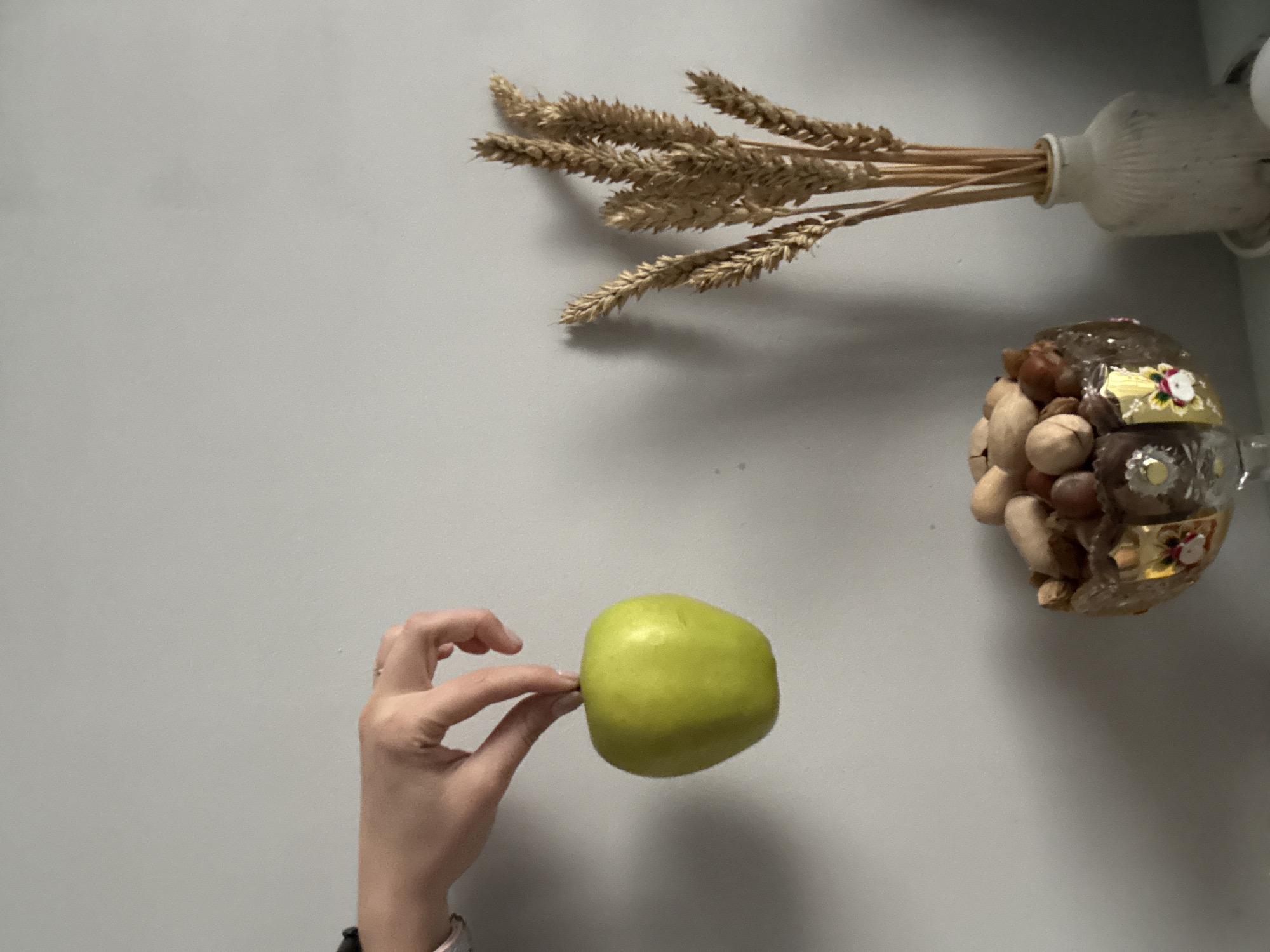A gray wall serves as the backdrop for a still life composition. In the upper right corner, dried wheat stalks are arranged in a small, white vase. Adjacent to this arrangement, a glass jar containing an assortment of nuts is positioned. In the lower left, a hand holds a green apple between the thumb and forefinger. No other details are visible regarding the location or city.