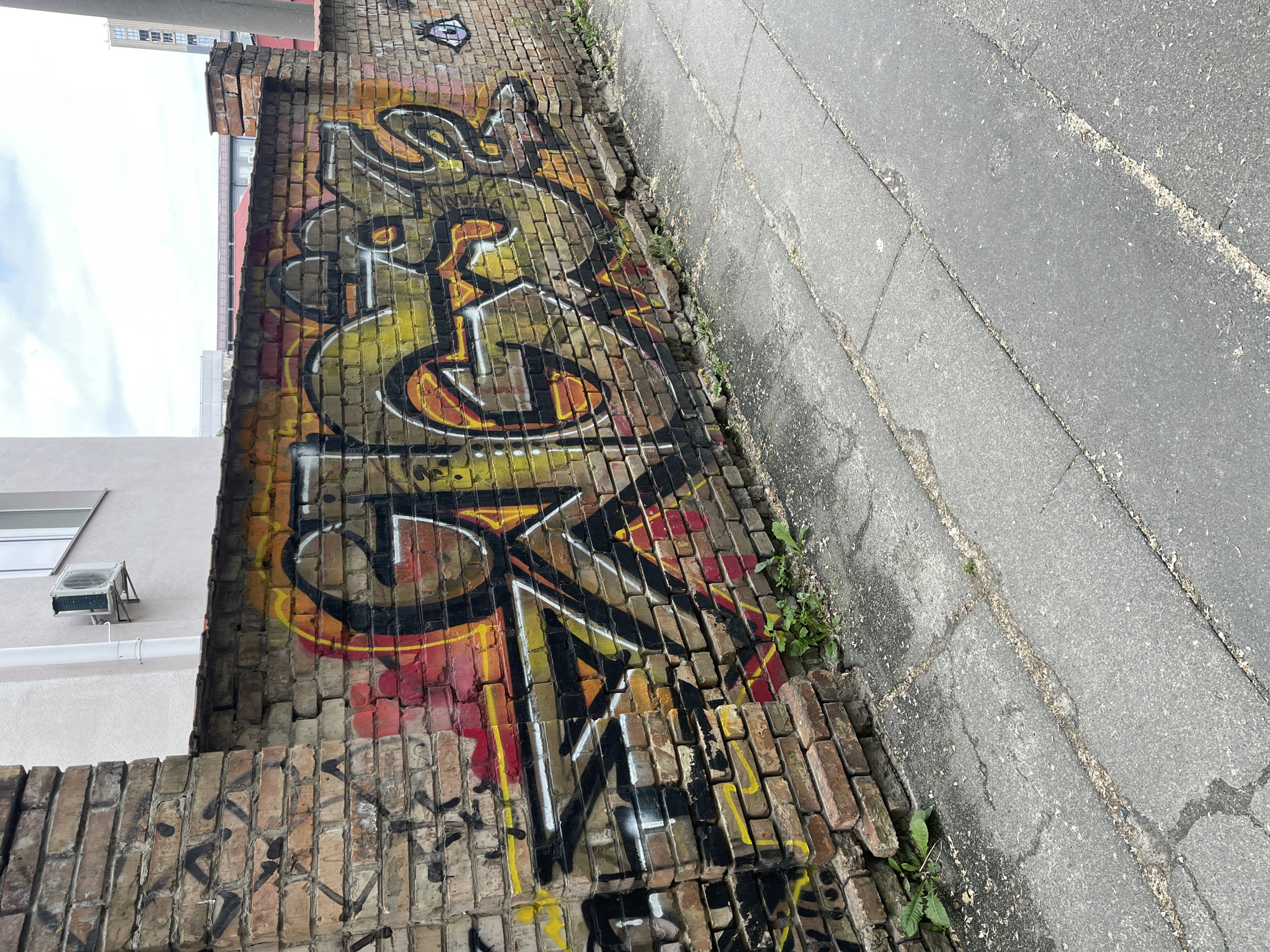 The image presents an outdoor scene dominated by a weathered, graffiti-covered brick wall running alongside a paved surface. The perspective is tilted, with the wall diagonally traversing the left side of the frame and the paved ground occupying the right.
The brick wall is constructed from reddish-brown bricks, many showing signs of age, chipping, and disrepair. A large, intricate piece of graffiti covers a significant portion of this wall. It features bold, stylized letters filled with bright yellow, orange, and red, outlined in stark black and white. While highly abstract and difficult to read definitively, elements resembling "S," "G," "E," "R," and "K" can be perceived within the design. Below this main artwork, and in other areas, are additional, smaller, black graffiti tags and scribbles that are illegible. A small, stylized graffiti eye is also visible near the top right edge of the wall.
To the right of the wall, a paved surface, likely a sidewalk or path, appears worn with cracks and varying textures of grey asphalt and concrete slabs. Small weeds and green plants sprout from the cracks in the pavement and along the base of the brick wall, indicating a somewhat neglected urban environment.
In the background, above the wall, glimpses of buildings are visible against an overcast, cloudy sky. On the left, a light-colored building with a window and an external air conditioning unit is partially seen. Further back and to the right, a taller structure with a reddish roofline can be discerned. The scene is illuminated by the diffused light of a daytime sky, suggesting either an overcast day or early/late daylight hours. The overall impression is one of a gritty, urban landscape.