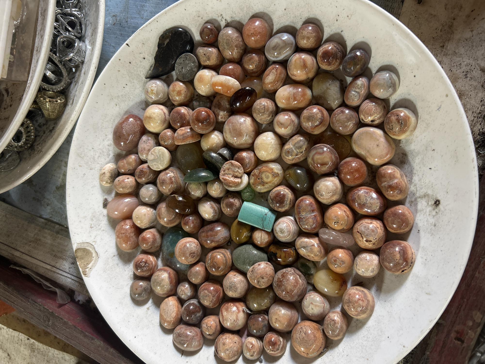 A white ceramic plate is filled with a collection of small, smooth, tumbled stones. The stones vary in color, with many exhibiting shades of reddish-brown, cream, and pink, often with swirling patterns or bands. Some stones are more opaque, while others are semi-translucent. There are also a few larger, darker, irregularly shaped stones, and one small, rectangular, turquoise-colored stone. The plate is slightly dirty with visible speckles. In the background, out of focus, is another plate or bowl containing various rings and jewelry, as well as some wooden elements. The lighting suggests an indoor setting, possibly a shop or a personal collection display, with a natural light source casting soft shadows. The scene appears to be located on a table or surface that is blue and worn. The image does not contain any identifiable elements that would suggest a specific location in Denpasar, Indonesia.