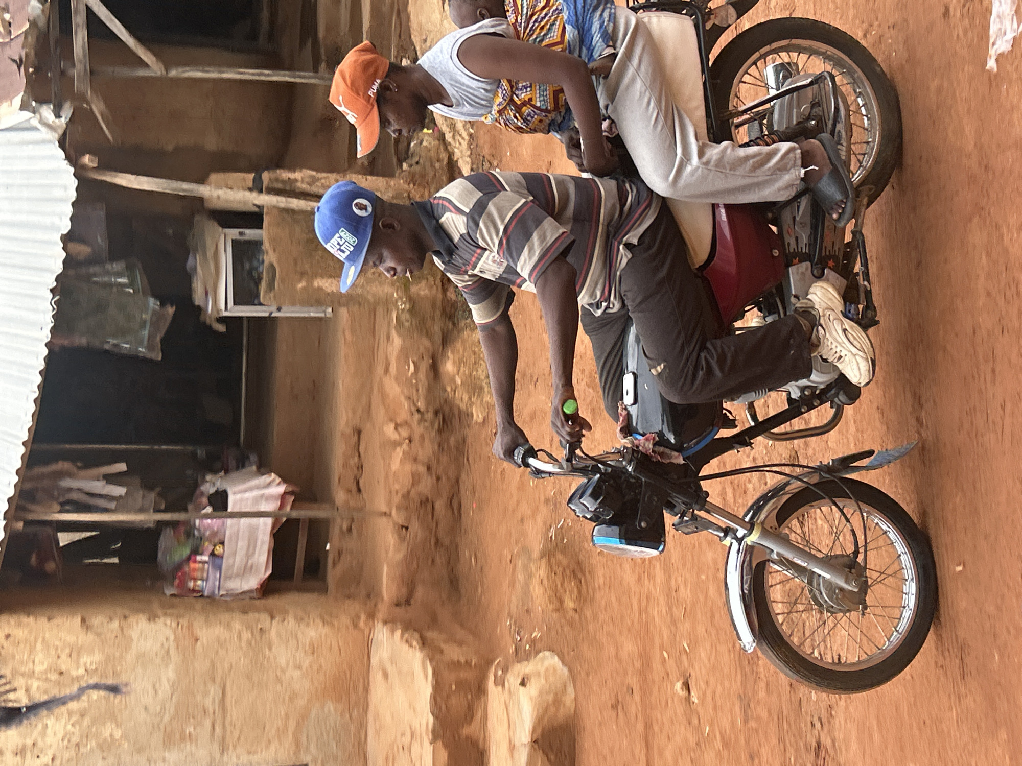 In Jos, Nigeria, two individuals are riding a motorbike on a dirt road. The driver wears a blue baseball cap, a striped shirt, and dark pants, while the passenger, perched behind, sports an orange baseball cap, a white top, and light-colored pants. The motorbike's design is simple, with a visible headlight and standard tires. The surrounding environment includes weathered buildings and a dirt surface.