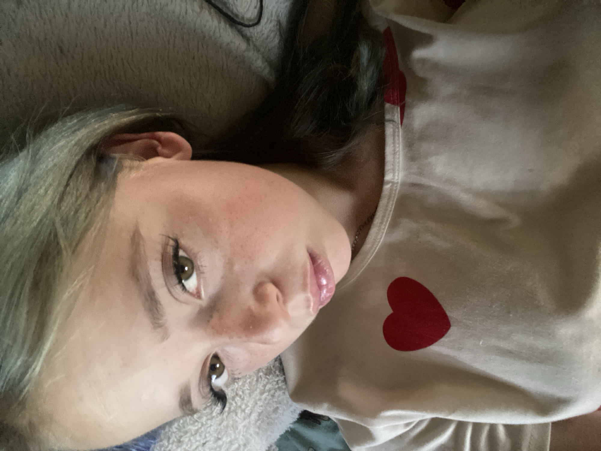 This media file is a close-up, sideways portrait of a young person lying down. The subject has light skin with freckles, prominent eyelashes, and light brown eyes. Their hair is light blonde with some greenish-blue highlights. They are wearing a light-colored shirt with two red heart designs on it, one near the collar and another on the chest. The person appears to be relaxed, with their head tilted and eyes looking slightly upwards. The background is blurred, showing a soft, textured surface that might be a blanket or pillow, and a hint of dark fabric. The lighting is soft and diffused, suggesting an indoor setting. There is no visible text in the image. The overall mood is intimate and candid.