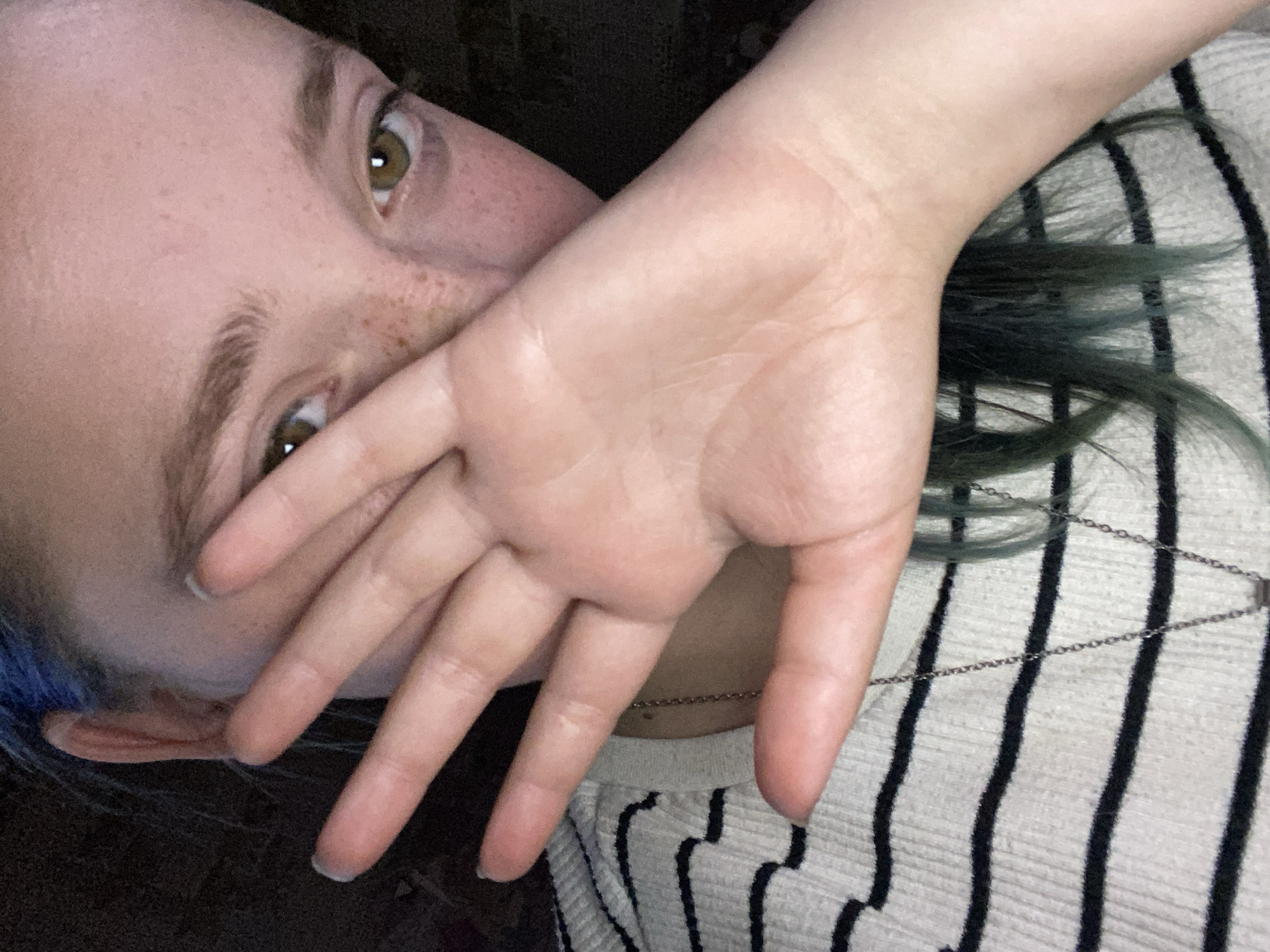 A close-up, head-and-shoulders shot shows a young person with blue-tinted hair and fair skin dotted with freckles. Their right hand is held up, obscuring the lower half of their face, with fingers spread. The person is wearing a black and white striped sweater and a silver necklace. The background is dark and indistinct, suggesting an indoor setting with low lighting. The scene does not provide any specific information about the city of Nova Praha, Ukraine.