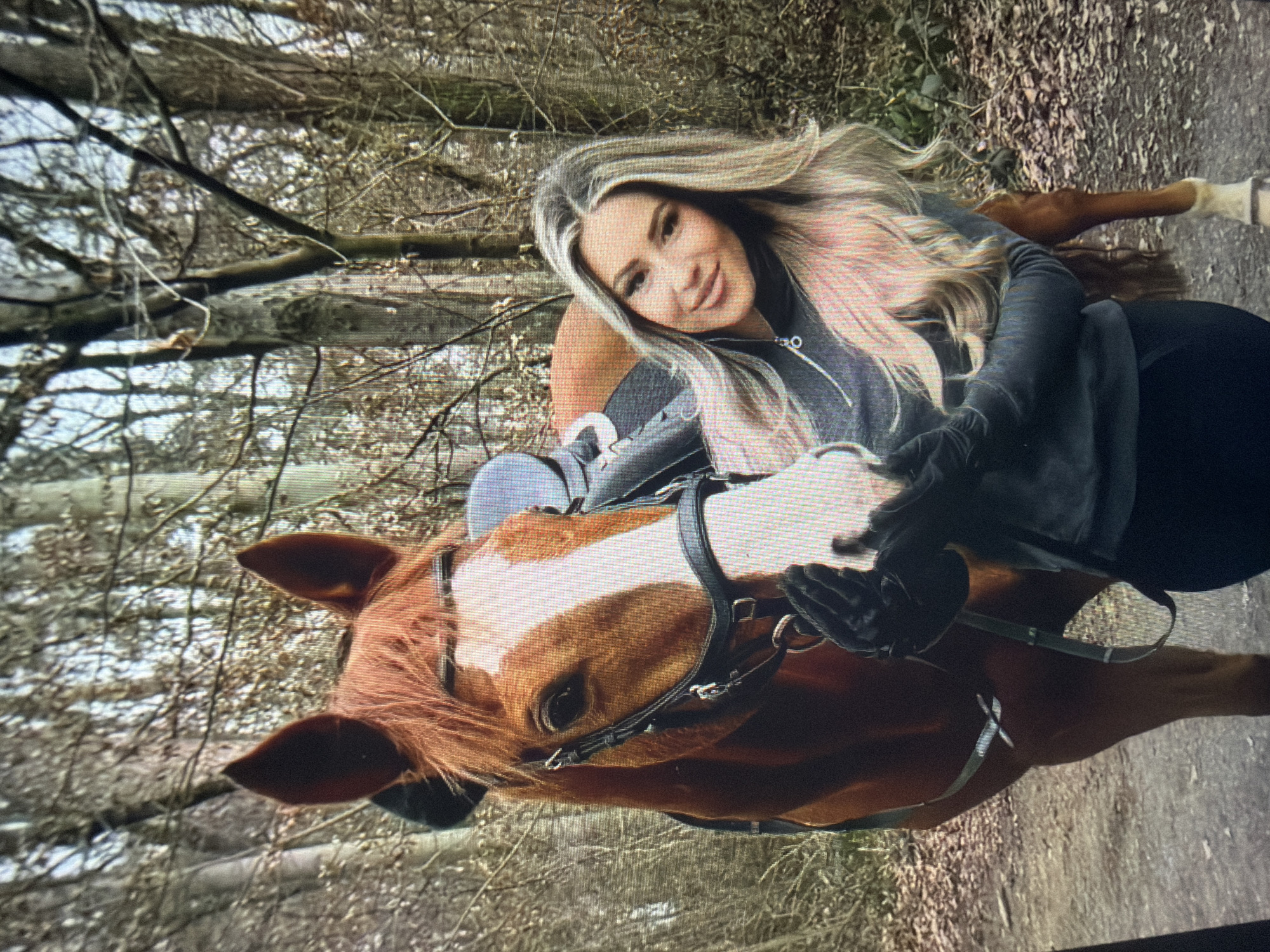 The scene, set in a wooded area in the city of Shumen, Bulgaria, features a woman and a horse standing on a dirt path.
The woman, positioned on the right side of the frame, has long, blonde hair with visible darker roots, parted on the left. She is smiling and looking towards the viewer. Her makeup includes dark eyeliner, mascara, and a light lip color. She wears a dark grey or black long-sleeved top with a metal zipper pull at the collar and dark pants. Her hands are covered by black gloves, and she holds the horse's muzzle with both hands.
The horse, a chestnut or bay in color with a prominent white blaze down its face, is equipped with a dark leather bridle. Its mane is a light brown-red. A dark saddle is visible on its back. The horse's left eye is visible, appearing dark and calm. Its ears are upright. The horse stands to the left of the woman, with its head slightly lowered, interacting with the woman's hands.
The background consists of a dense forest with numerous bare deciduous trees and branches, indicating a colder season such as late autumn or winter. The ground beneath the subjects is a mix of dirt, fallen leaves, and sparse vegetation. The overall lighting suggests an overcast or shaded outdoor environment.