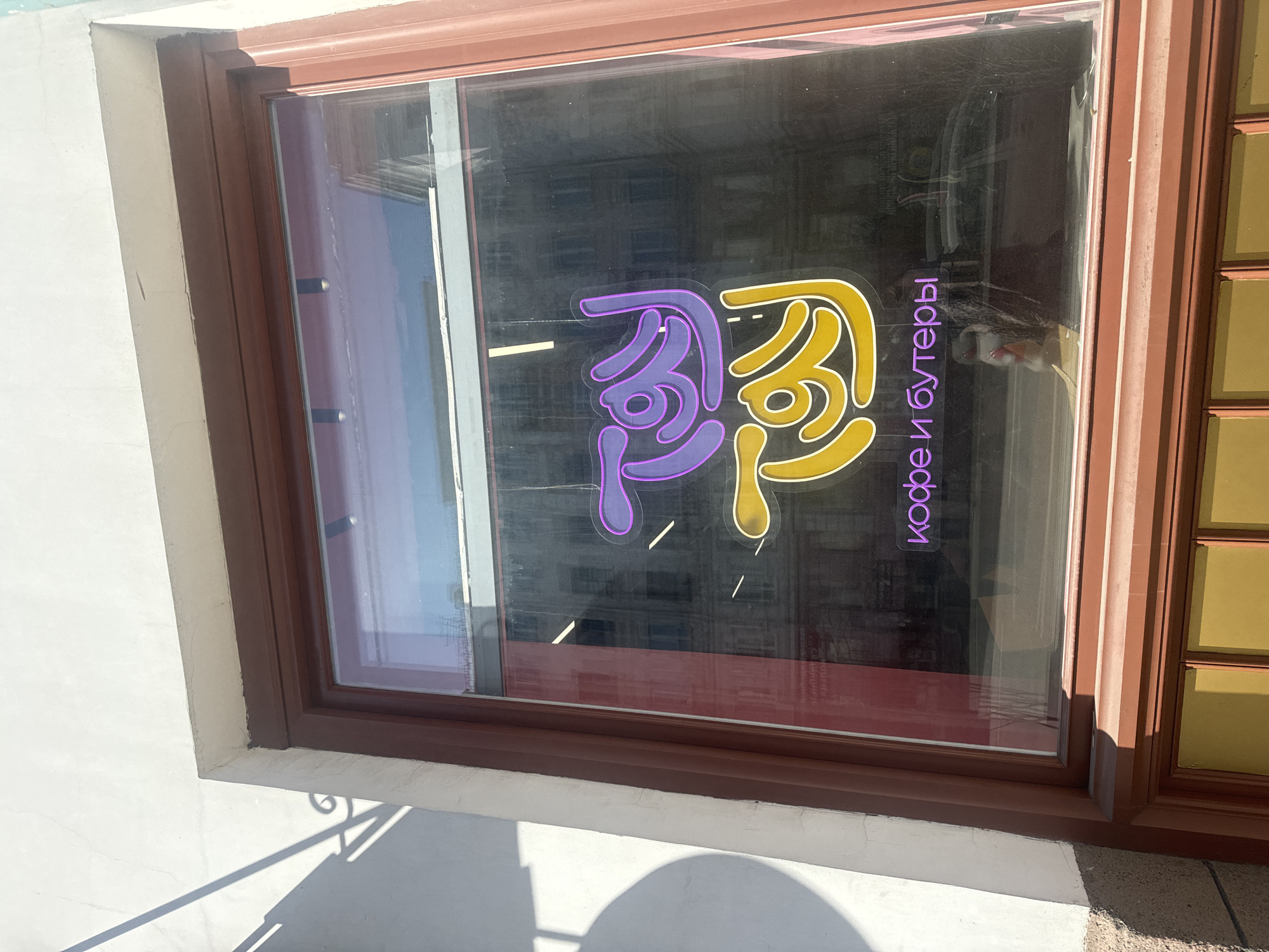 A sunlit shop window, part of a building facade, is captured in a close-up, likely at ground level, in Krasnoyarsk, Russia. The window features a sturdy brown frame with decorative molding. The building's exterior wall, visible on the left and below the window, is white plaster, showing some minor cracks.
Inside the window, a vibrant, stylized sign, appearing to be made of neon or backlit plastic, is prominently displayed. It features two abstract, curvilinear shapes: one purple and one yellow, both outlined in white, serving as a distinctive logo. To the right of these shapes, purple vertical text, also in a neon style, reads "кофе и бутеры." This translates from Russian to "coffee and sandwiches" or "coffee and butties," indicating the type of establishment.
The window glass reflects indistinct architectural elements, possibly from buildings opposite or objects within the shop. On the far right, a section of an adjacent window with smaller, yellow-tinted panes is visible. Bright sunlight casts sharp shadows on the white wall and pavement, suggesting a clear, sunny day. No people or direct activity are visible, but the shop sign implies a commercial setting.