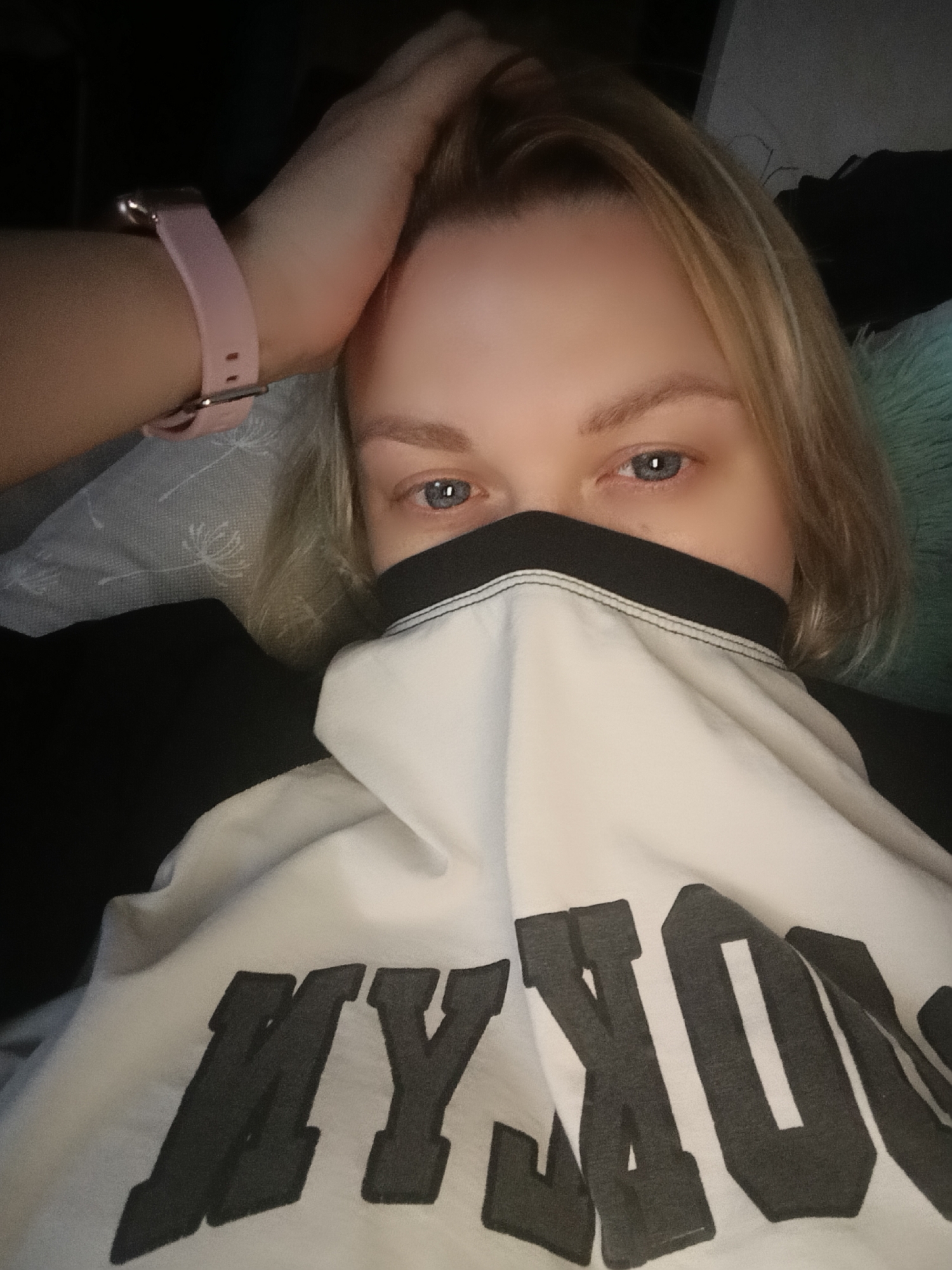 A person with light blonde hair and blue eyes is lying down, partially obscuring their face with a white t-shirt that has black lettering. The lettering appears to spell "MYKO...," but is cut off by the frame. The person is wearing a pink smartwatch on their left wrist. The background is dark, with a glimpse of a light-colored pillow.
There is no visual evidence in the provided image to suggest any link to the city of Chaikovsky, Russia. The image shows an individual inside a dimly lit location.