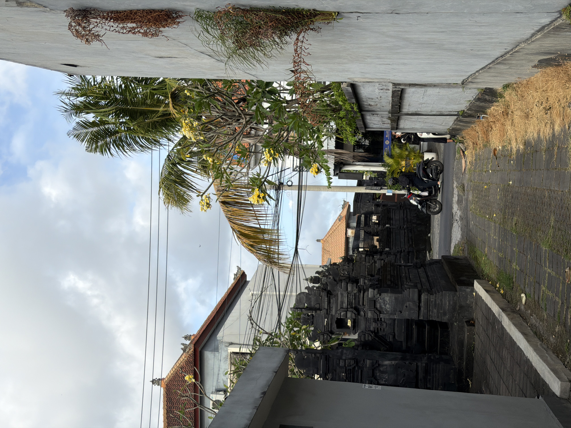The image shows a street scene in North Kuta, Indonesia. The street is narrow and paved with dark gray bricks. On the right side of the street is a concrete curb and a sidewalk made of the same dark gray bricks. A building with a red tile roof is visible on the left side of the street. A large, dark gray stone structure with multiple tiers stands next to the building. A palm tree and other trees with yellow flowers grow between the buildings and the street. Overhead, there are several power lines. A motorcycle with two people on it is parked on the right side of the street in front of a building with a blue and white sign. The sky is overcast with gray clouds. Some dried vegetation hangs from the top of the building on the right.