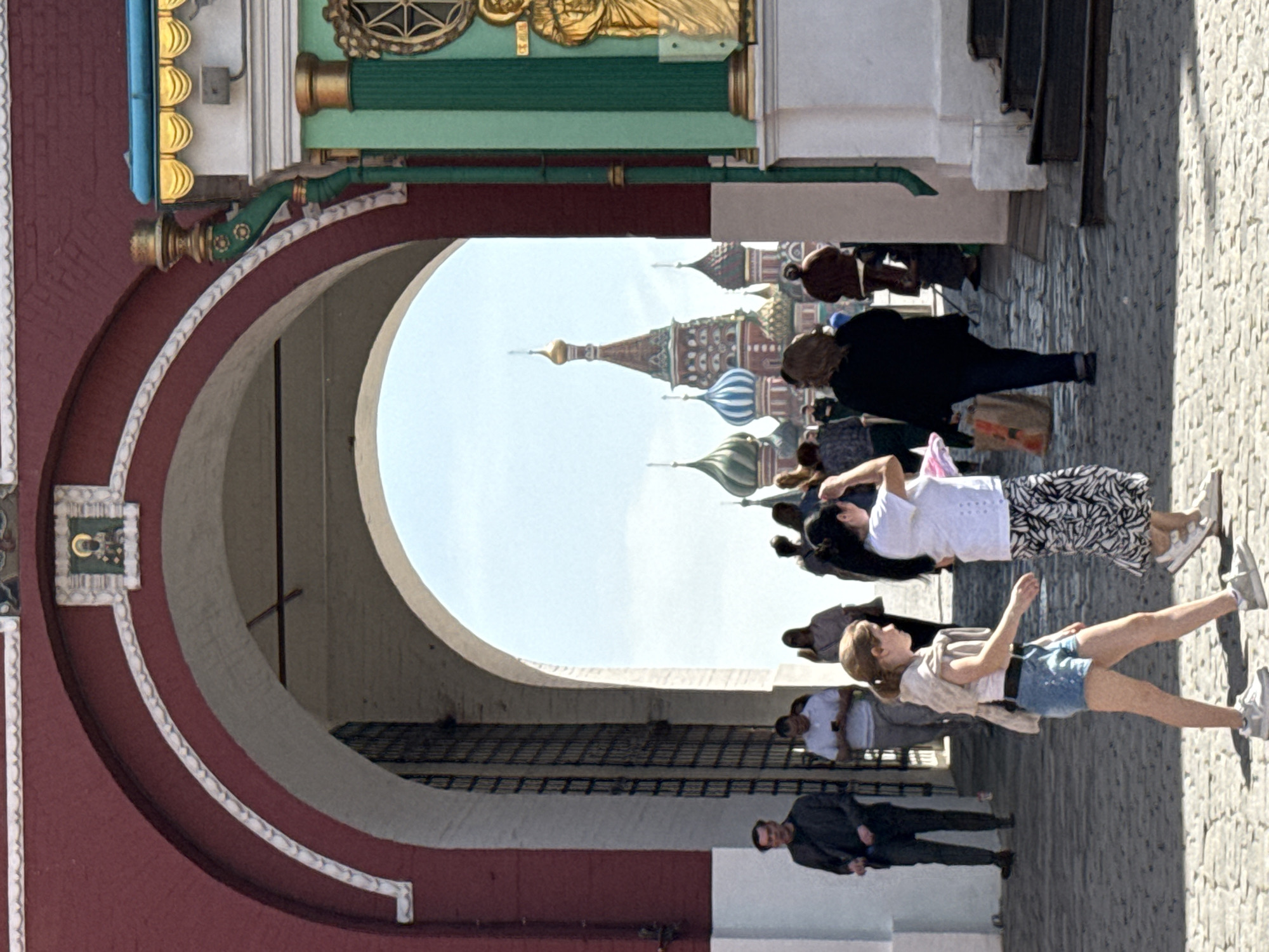 View through an archway, with St. Basil’s Cathedral visible in the background. Several people are walking, standing, or sitting in the foreground. The ground appears to be cobblestone.
