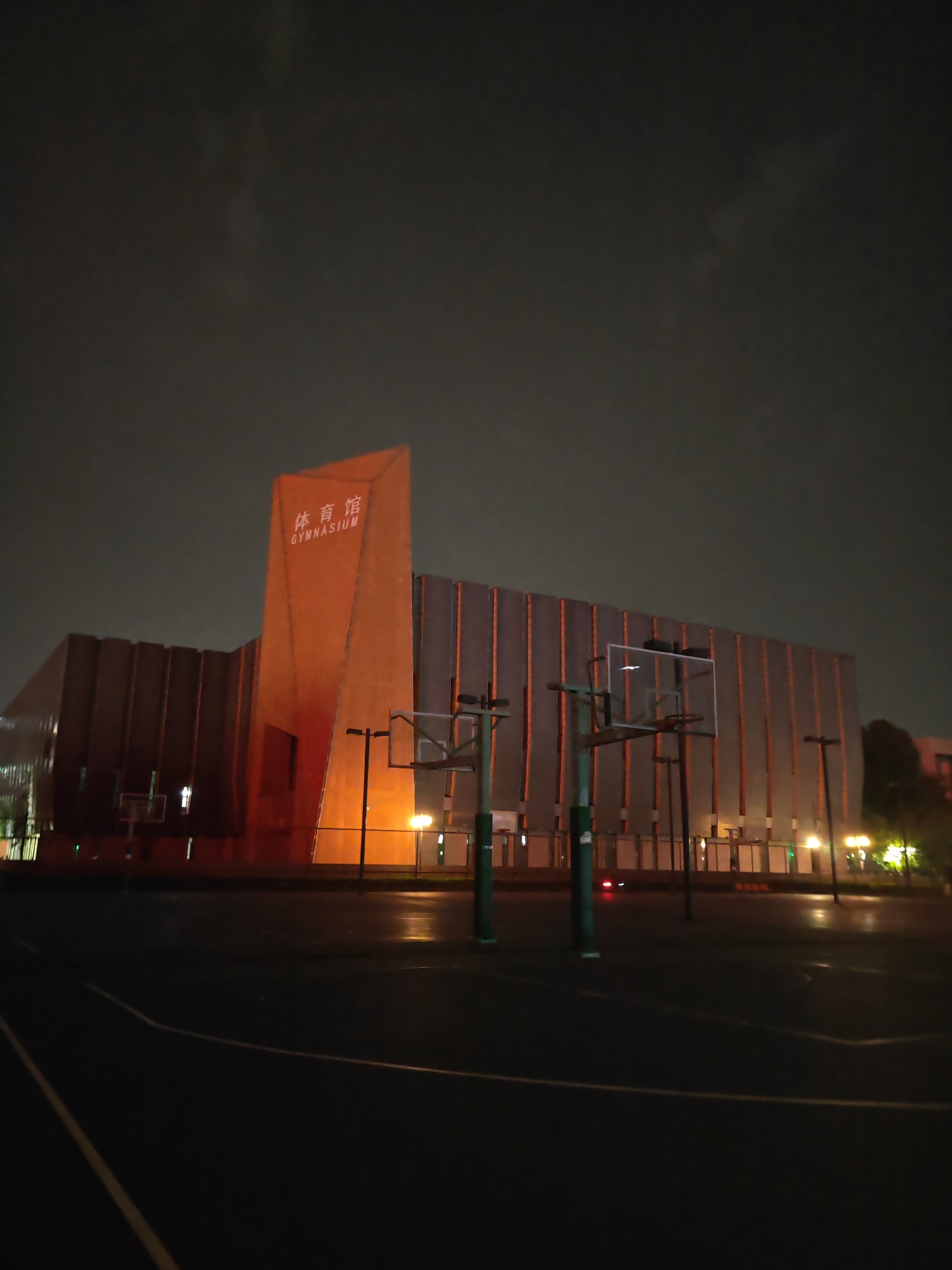 The image depicts an exterior view of a gymnasium at night, located in Jiangning District, China. The gymnasium's facade features a prominent orange tower with white lettering "体育馆 GYMNASIUM." To the right is a dark rectangular building with vertical lines. In front of the gymnasium, there is a basketball court with several hoops visible, along with the lines drawn on the court. The lighting is subdued, with a mix of orange light emanating from the building and the yellow from the street lamps. The sky is dark and overcast. No people are visible in the scene.