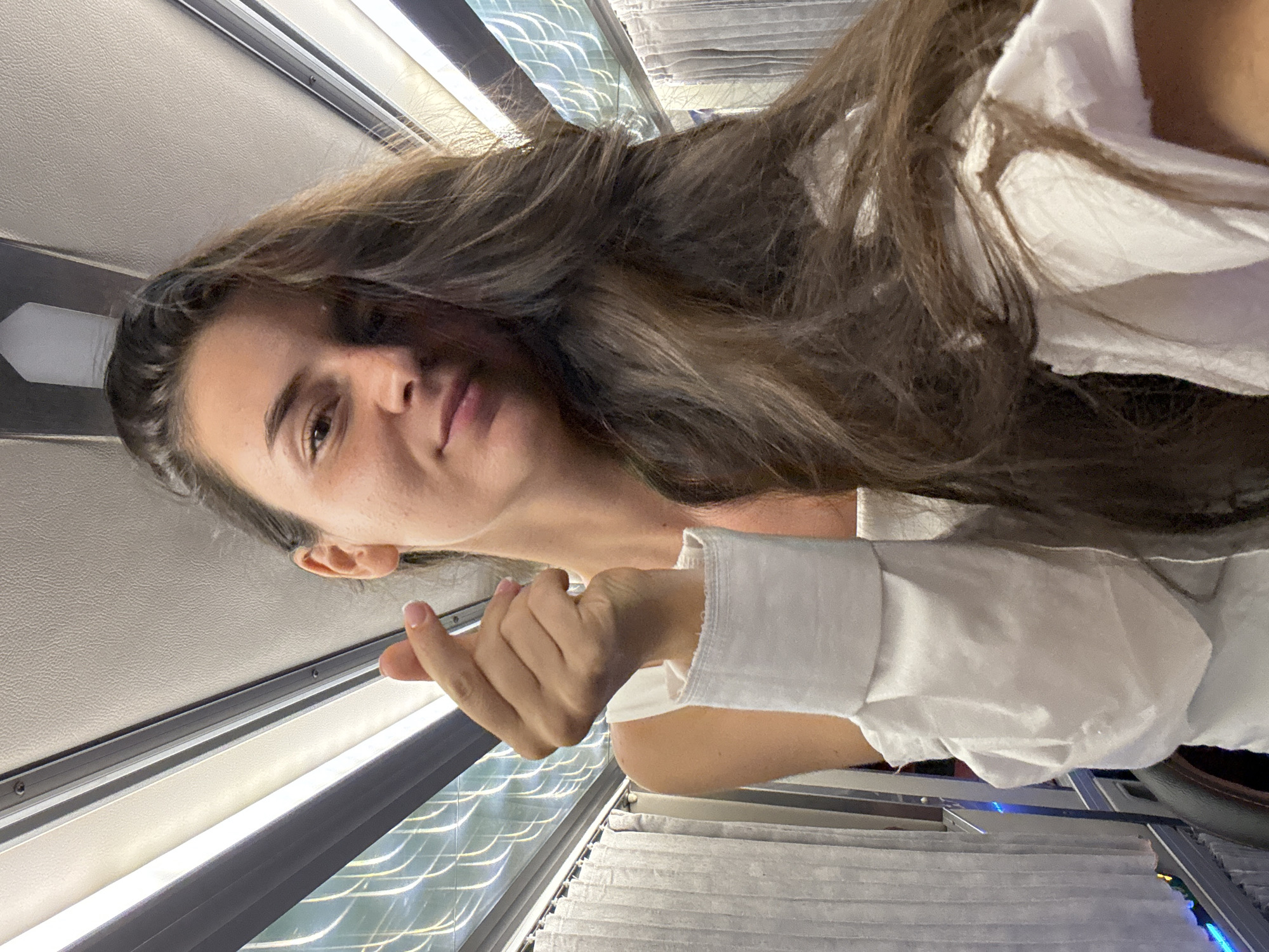 A woman with long brown hair, wearing a white shirt with the sleeves rolled up, is sitting by a window on what appears to be a bus or train. She is looking at the camera with a neutral expression, and her hand is held up near the window. The window has drawn blinds. The location is indicated as Phu Thuy Ward, Vietnam, but there is no visible evidence of this in the image.