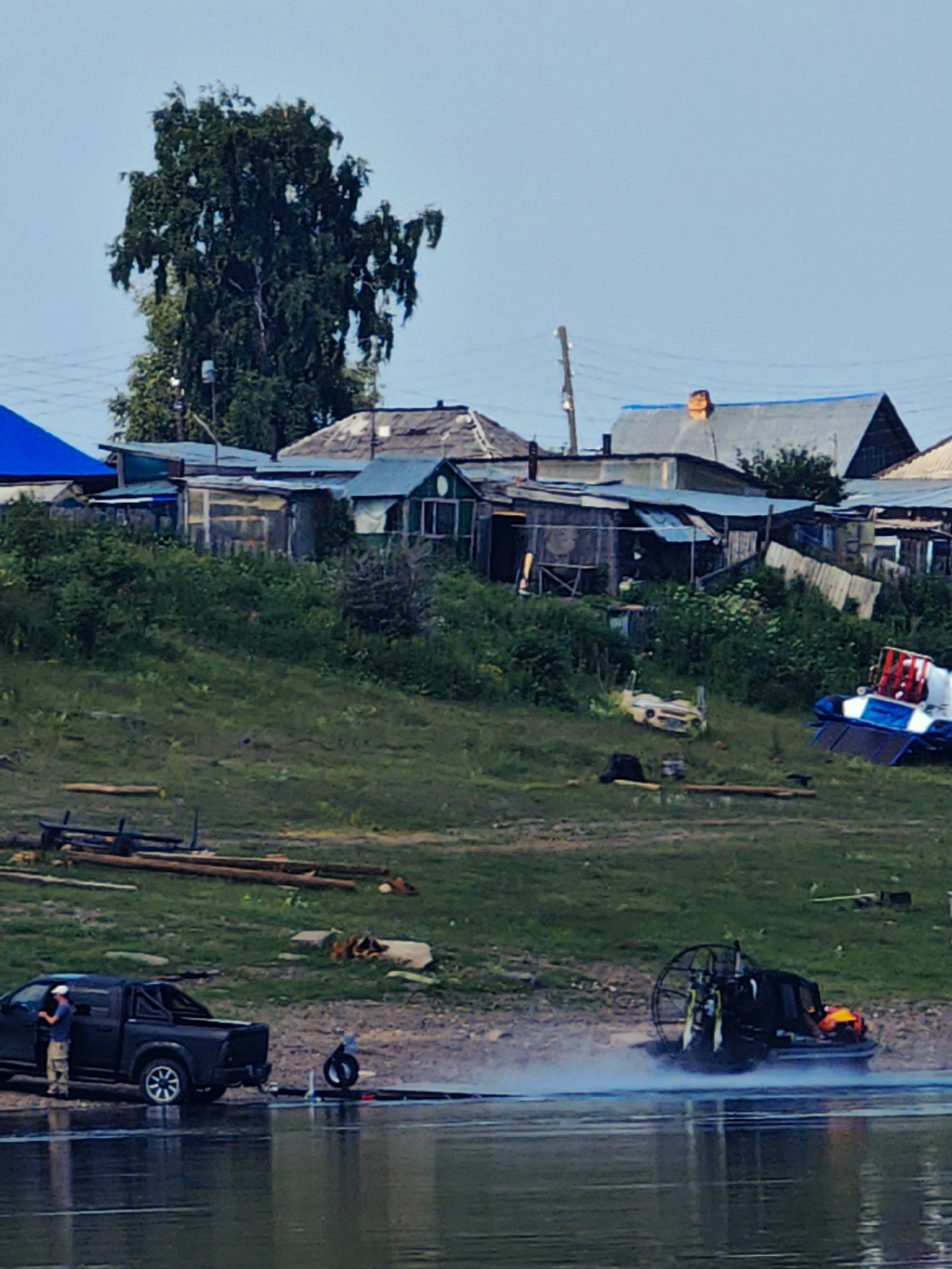 The image depicts a waterfront scene in the village of Pogodaevo, Russia, on what appears to be a clear day. In the foreground, the calm, reflective surface of a body of water dominates the lower portion of the frame.
On the shore to the left, a dark-colored pickup truck is parked, with a person wearing a white cap and light-colored pants standing near the open driver's side door. Adjacent to the truck, a tire lies on the ground next to a water-level launch area. Further out on the water, a black hovercraft is in motion, emitting a plume of mist and spray as it travels.
The shore itself is a grassy slope, with various objects scattered on it. To the left, a pile of logs and wooden planks are visible. Further up the slope, a collection of various structures are situated, appearing to be a mix of small houses, sheds, and outbuildings. These buildings are varied in construction, with some featuring tin or corrugated metal roofs, and others appearing to be more rustic wooden structures. A tall, leafy tree stands prominently behind these buildings.
In the mid-ground and background, the landscape continues with more greenery and the structures of the village. Power lines and a wooden utility pole are visible in the upper portion of the image. The sky is a pale blue, with no clouds apparent. The overall impression is of a functional, somewhat rural, waterside settlement.