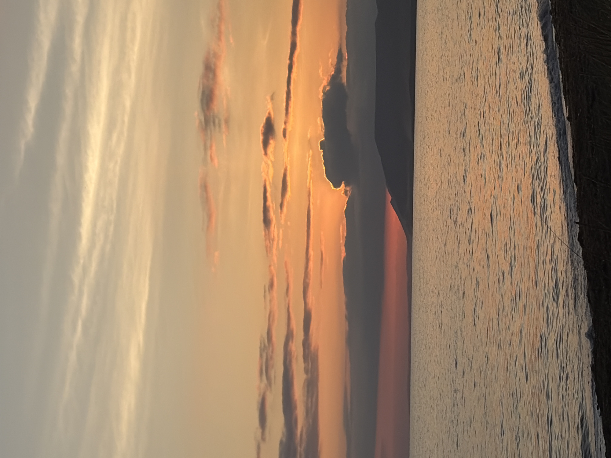 The image depicts a serene natural landscape at sunset. The sky is filled with a gradient of warm colors, ranging from soft yellow and orange near the horizon to a muted grey and pink higher up. Thin, wispy clouds are scattered across the sky, catching the fading sunlight.
Below the sky, a dark silhouette of a distant mountain range stretches across the horizon. The sea occupies the middle ground, its surface reflecting the colors of the sky with gentle ripples. The water is calm, with a hint of pink and orange hues washing over it.
The foreground shows a dark, indistinct shoreline. Some dry grasses or reeds are visible along the edge of the water. The overall impression is one of tranquility and natural beauty, characteristic of a coastal sunset. The location is identified as Ozerskoe, Russia.