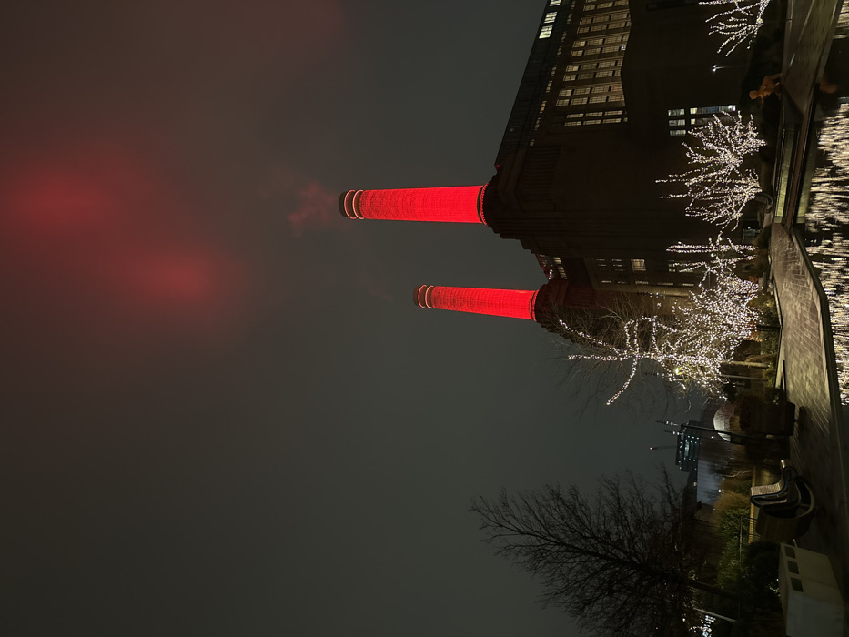 Battersea Power Station at night is lit with red lights on its chimneys. The iconic structure is mirrored by the water below. Trees with twinkling white lights line the waterfront near the power station. The sky is overcast, creating a dramatic and atmospheric backdrop to the scene. Bare tree branches are visible in the foreground.