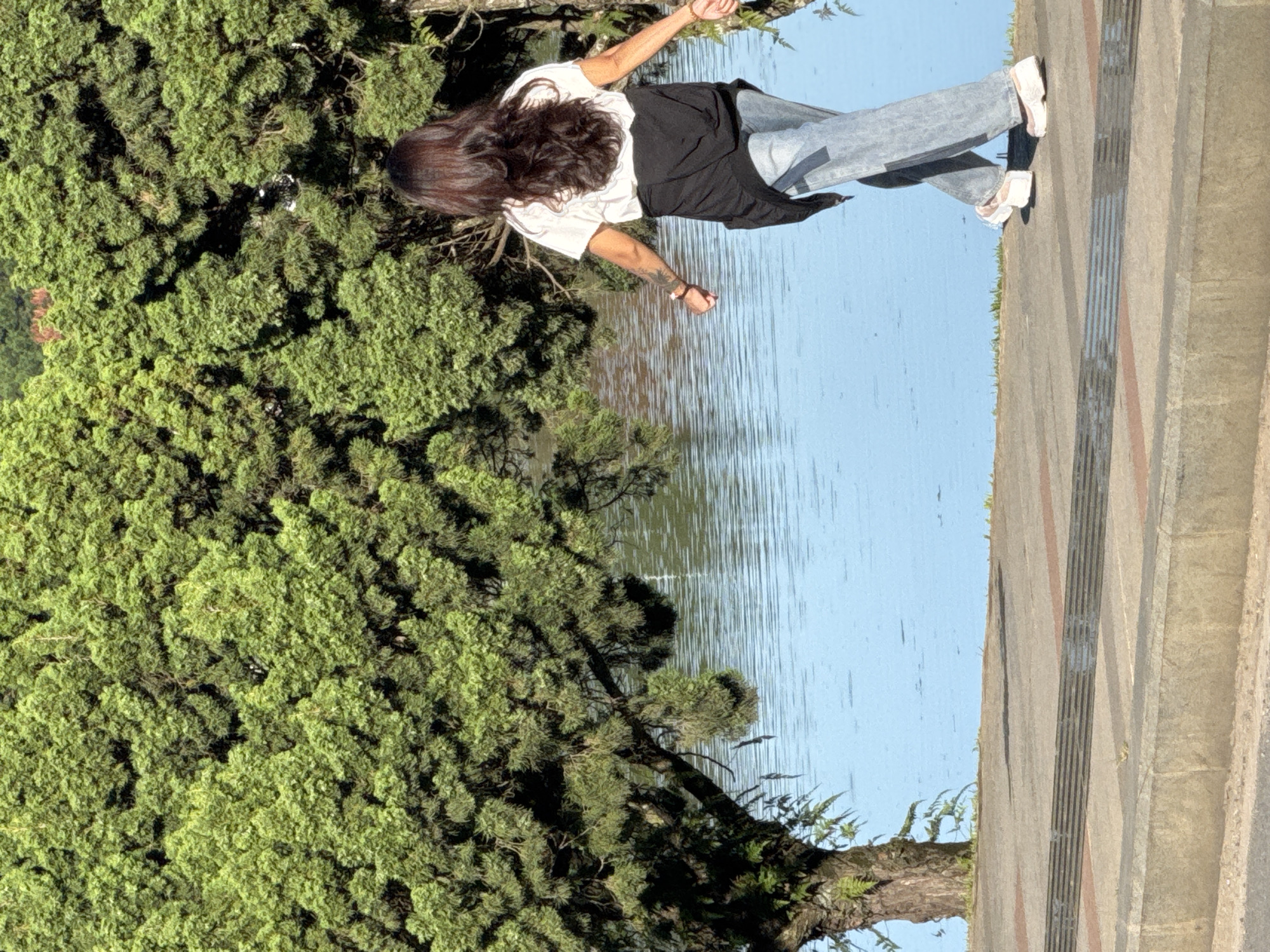 A woman with long, dark hair, wearing a white t-shirt and blue jeans, walks along a paved pathway next to a body of water. A black jacket is tied around her waist. The pathway has a textured surface with contrasting colored stripes. Behind the pathway is a large body of calm, blue water reflecting the sky. To the left of the woman and the water is a large, dense green tree with a thick trunk. The scene appears to be in a park or recreational area, likely Xuan Huong Lake in Da Lat, Vietnam, given the lush greenery and the presence of a large lake common in the city. The lighting suggests it is daytime, with the sun casting shadows on the pathway.