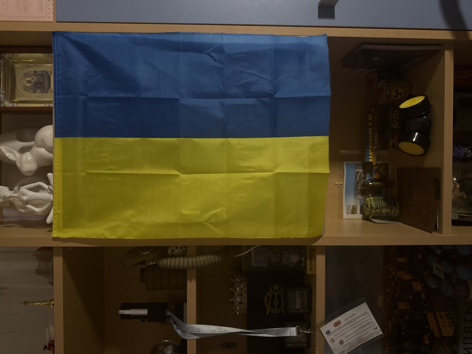 A Ukrainian flag hangs on a shelf with various objects. A framed picture and white figurines are to the left. A stack of items, including a magazine and cylindrical canisters, is on the right. Below the flag, another shelf contains a gas mask, a gun, and a card with information. The shelves are wood-toned.