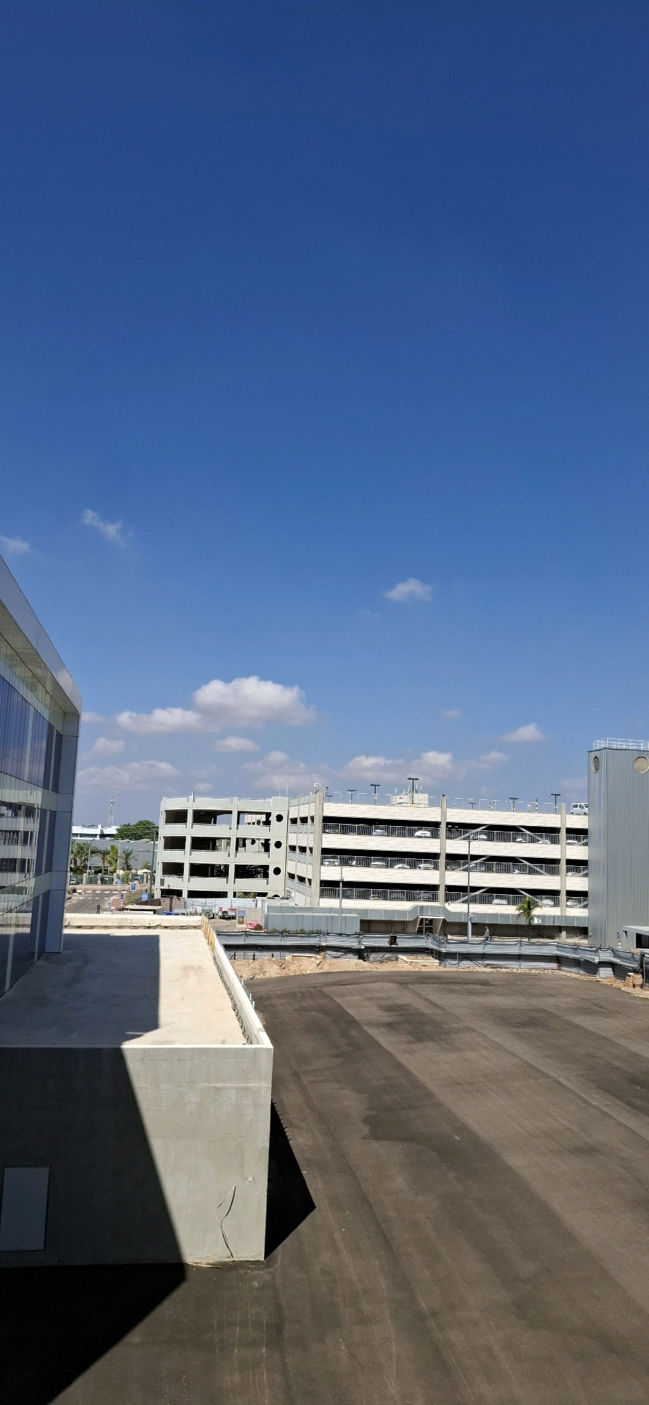 The image captures a view of a construction site or recently developed area, likely part of an industrial or commercial zone, in Kiryat Gat, Israel. The foreground features a portion of a concrete structure, casting a distinct shadow on a dark asphalt surface. In the mid-ground, a multi-level parking garage, constructed from concrete and metal railings, dominates the scene. This structure exhibits an unfinished or brutalist architectural style, with open floors and visible support columns. Some vehicles are visible on the upper levels of the parking garage. To the left, a modern building with glass facades is partially visible, suggesting a contrast between older and newer developments. Beyond the parking garage, trees and other low-rise structures are discernible, indicating the broader urban or suburban context. The sky above is a clear, vibrant blue with scattered white clouds, suggesting fair weather. The overall impression is one of ongoing development and infrastructure expansion within the city.