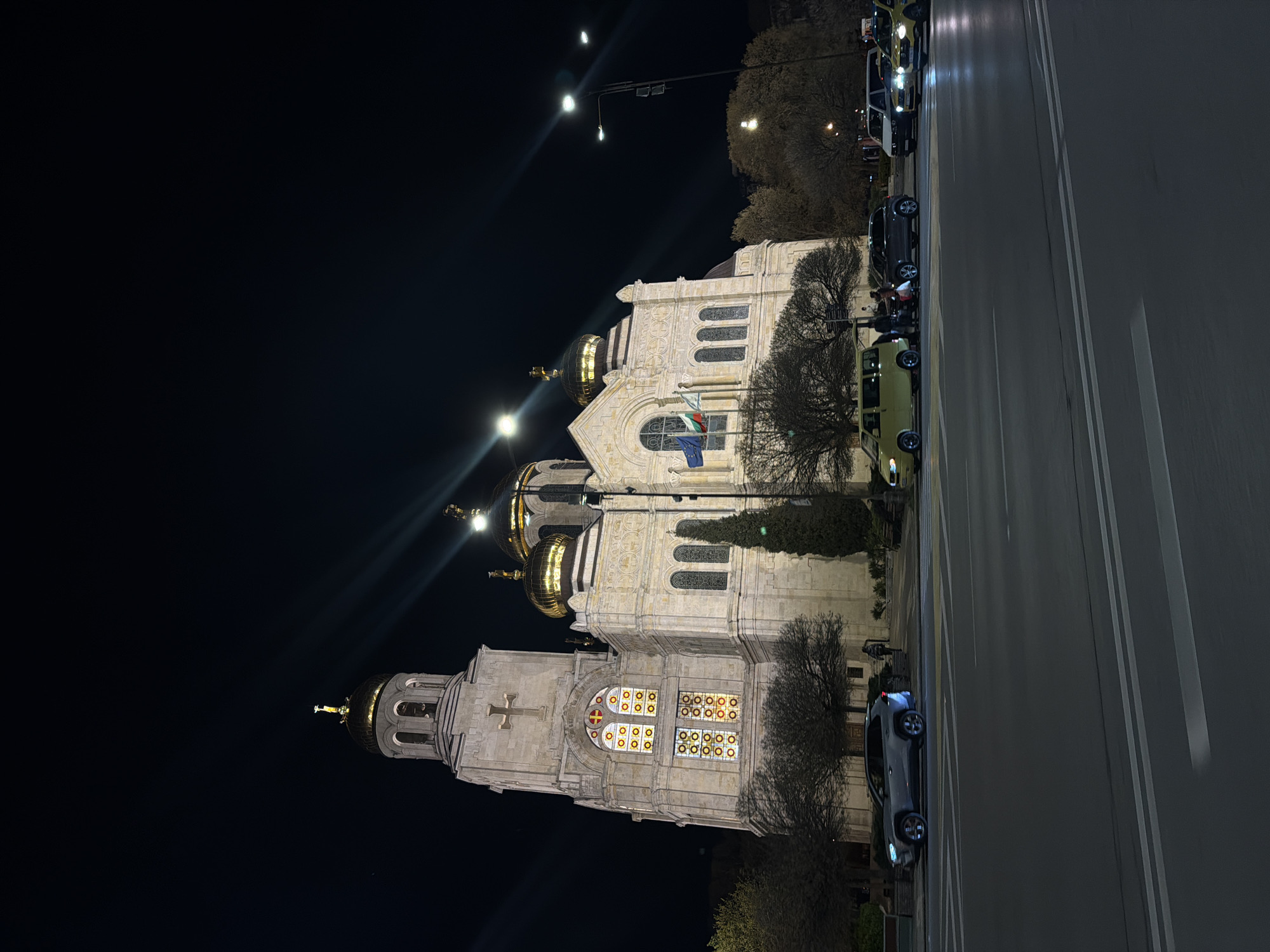 At night, the Dormition of the Mother of God Cathedral in Varna, Bulgaria, is illuminated, showcasing its golden domes and detailed stone facade. A Bulgarian flag hangs near the entrance. Cars are parked along the adjacent street, and street lights shine in the background. Bare trees surround the cathedral.