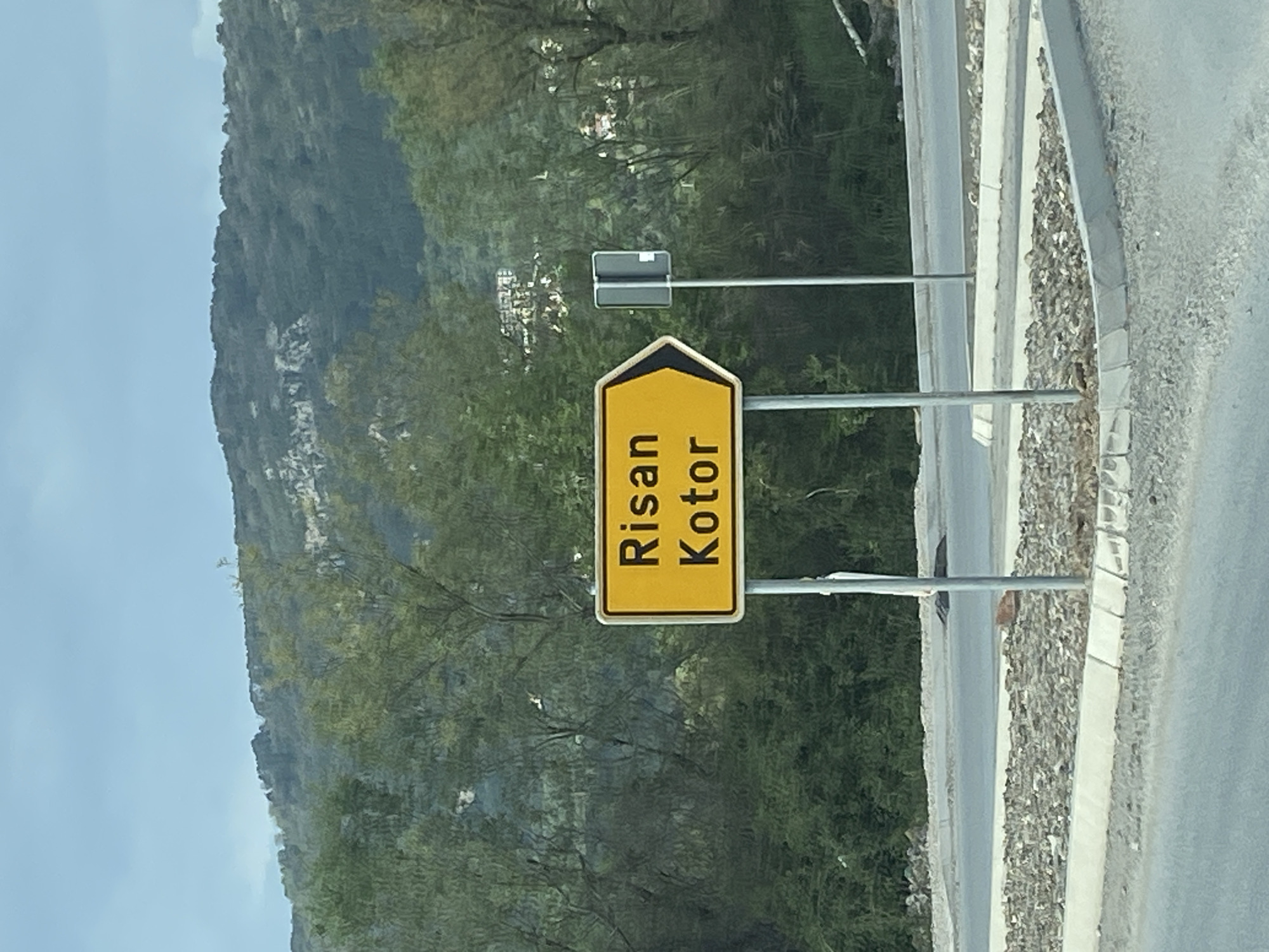 The image shows a yellow and black road sign indicating the directions to Risan and Kotor in Kavač, Montenegro. The sign is mounted on metal poles beside a road with a concrete barrier. Behind the sign is a hillside covered in green vegetation, and a blue sky is visible above. A smaller rectangular sign is situated above the direction sign. The photograph is taken from a roadside location.