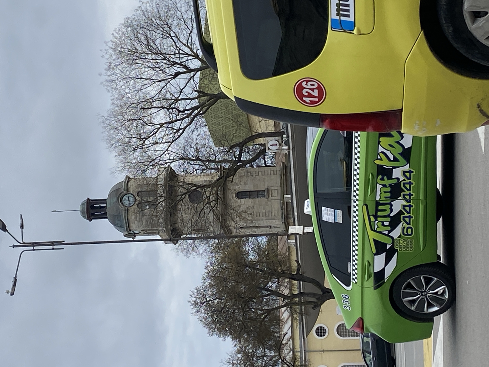 The image shows a street scene in Varna, Bulgaria. A yellow taxi with the number "126" is parked next to a green taxi bearing the name "Triumph Taxi" and the phone number "644444". The number "336" is also visible on the green taxi. In the background, a clock tower is visible, with leafless trees framing it. A street lamp with multiple lights stands to the left of the clock tower. The sky is overcast.