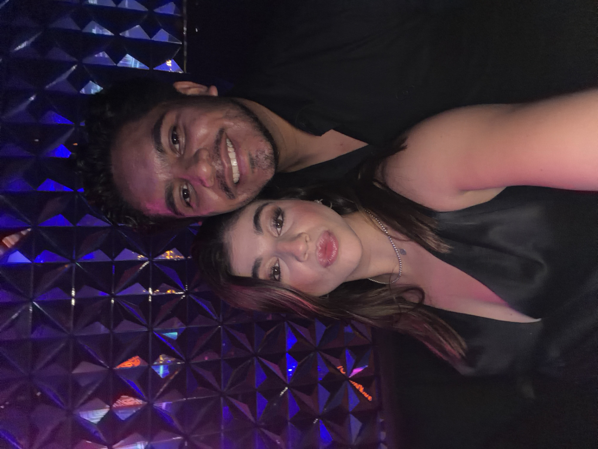 A selfie depicts a man and a woman indoors, close together. The man, on the left, has dark hair, a trimmed beard, and is smiling broadly. The woman, on the right, has long brown hair and is looking towards the camera. Both are wearing dark clothing. The background consists of a textured, geometric pattern with reflective, blue and purple lighting. It is not possible to verify if the image was taken in San Bernardino Tlaxcalancingo, Mexico.