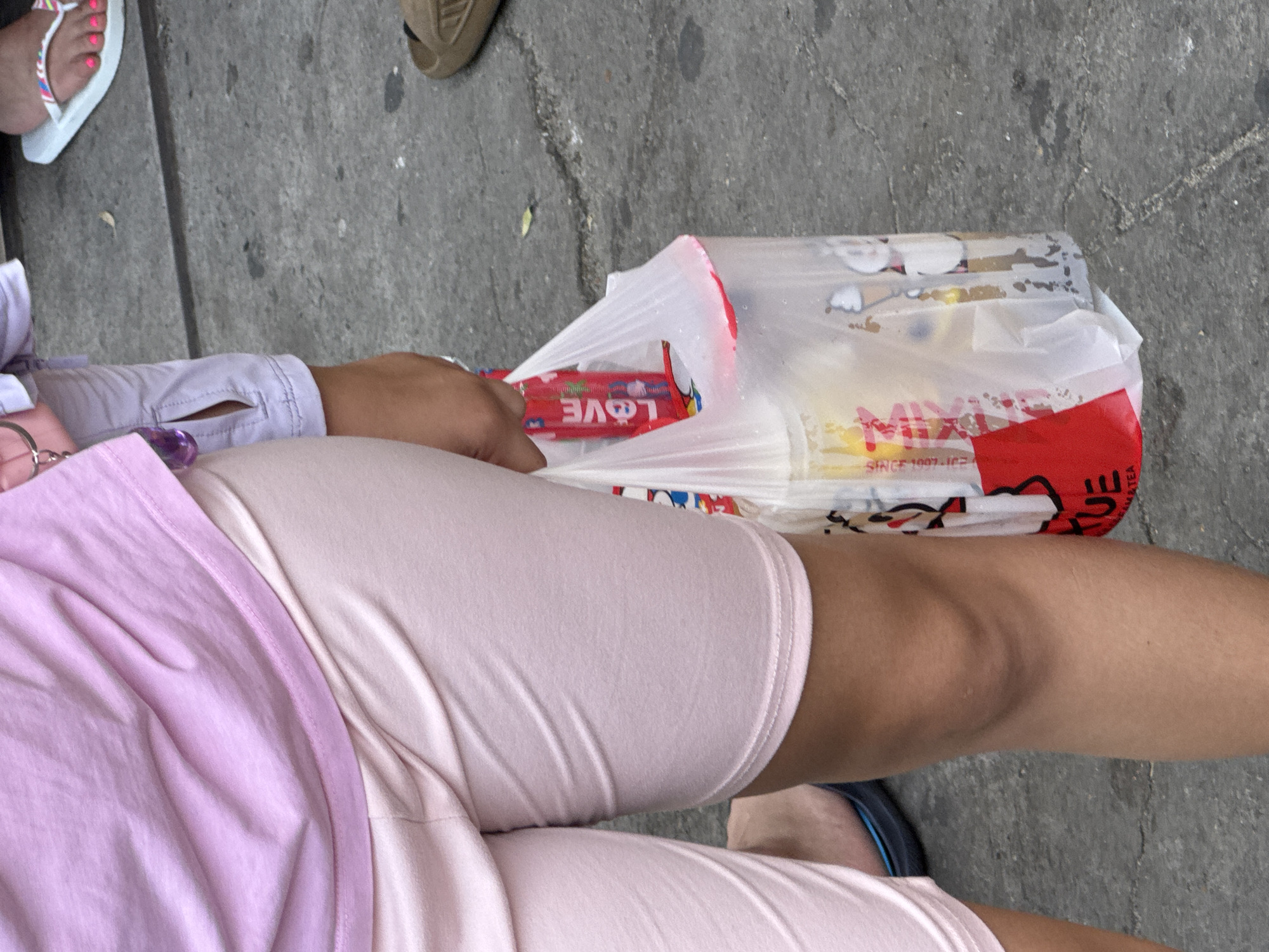 The image depicts an outdoor street scene in Nha Trang Ward, Vietnam. In the foreground, a person is visible from the upper thigh to the mid-calf, wearing a light pink short-sleeved shirt and light pink shorts. The person's right hand holds a clear plastic bag containing multiple items, including at least two beverages. One item inside the bag is a red container or package marked "L0VE" (with a heart symbol for 'o'). Another cup inside the bag features white cartoon illustrations, possibly of ice cream cones. A red label on the exterior of the plastic bag partially displays the text "UE" and "MATEA," indicative of a milk tea product. The ground consists of uneven grey concrete with visible cracks, minor scuff marks, and small dark specks of debris. In the background on the right, a person's feet are visible, wearing white flip-flops with multicolored straps, and the toenails are painted bright pink. A brown slide sandal is partially visible beneath the flip-flops. To the far left, a person wearing light purple shorts with a visible side pocket is partially seen, along with the edge of a dark blue sandal.