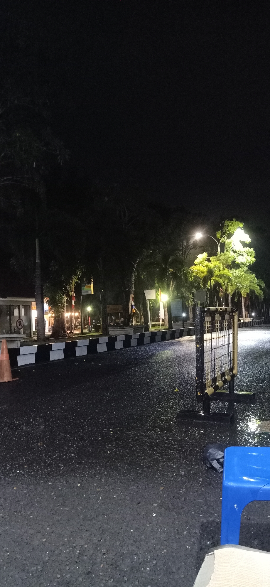 An exterior nighttime shot shows a wet, dark asphalt road in Tambontunong, Indonesia. The road is bordered by a black and white striped barrier on the left. Trees are visible in the background, some illuminated by streetlights, creating a patchy, green glow. On the right side of the road, a metal barrier with a grid pattern stands. A blue plastic chair and a black trash bag are on the road.