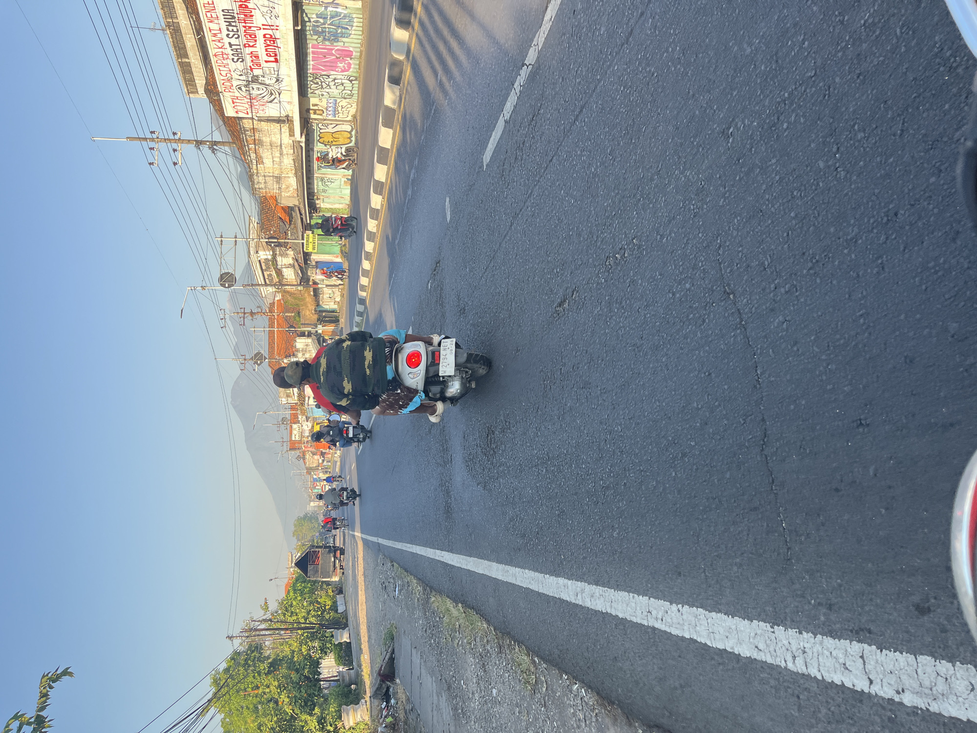 The image depicts a street scene in Gedang, Indonesia, with a clear blue sky and a distant mountain. The foreground shows the road surface with a white lane marker. In the midground, several motorcycles are visible, with one in the center carrying two people. The rider in front wears a black and red jacket, and the passenger behind wears a green and black camouflage jacket and patterned shorts. The motorcycle has a license plate that reads "W 2794 NET".
The street is lined with buildings on the right side. These buildings have tiled roofs and some feature graffiti. One building has a large banner with red text. On the left side of the road, there are trees, utility poles, and a parked truck. Several other motorcycles and a bicycle are also present on the road, indicating a moderately busy street. The overall impression is a sunny day in a developed area with ongoing traffic.