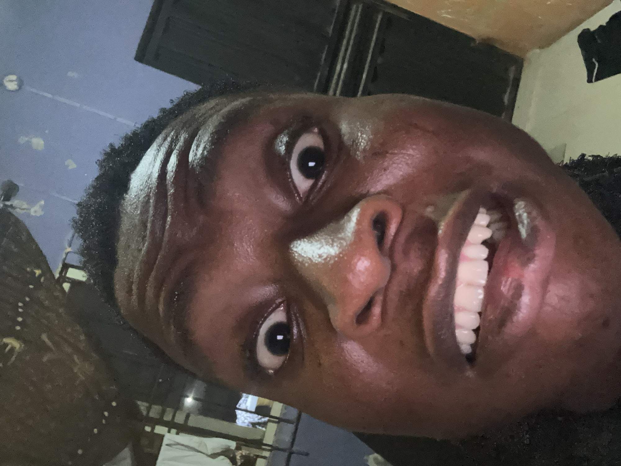 A close-up, tilted perspective image shows a Black male face with dark skin, shiny from sweat or moisture. His dark curly hair is visible along the hairline. The eyes are wide open, and the teeth are exposed in a wide smile or grimace. The background is blurry, showing hints of a room with possible window bars and a bed with white sheets. The location cannot be determined.
