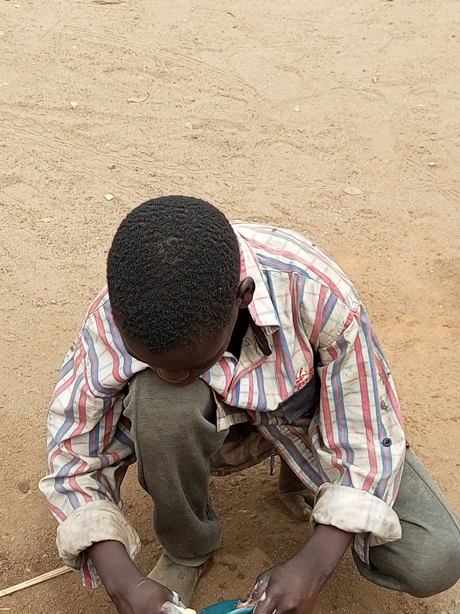 A young boy is crouching on the sandy ground in Gombe, Nigeria. He is wearing a striped long-sleeved shirt and grey pants. His hands are clasped together in front of him, and he appears to be examining something in his hands. The ground is made up of sand and small stones. The image is taken from a high angle, looking down at the boy.