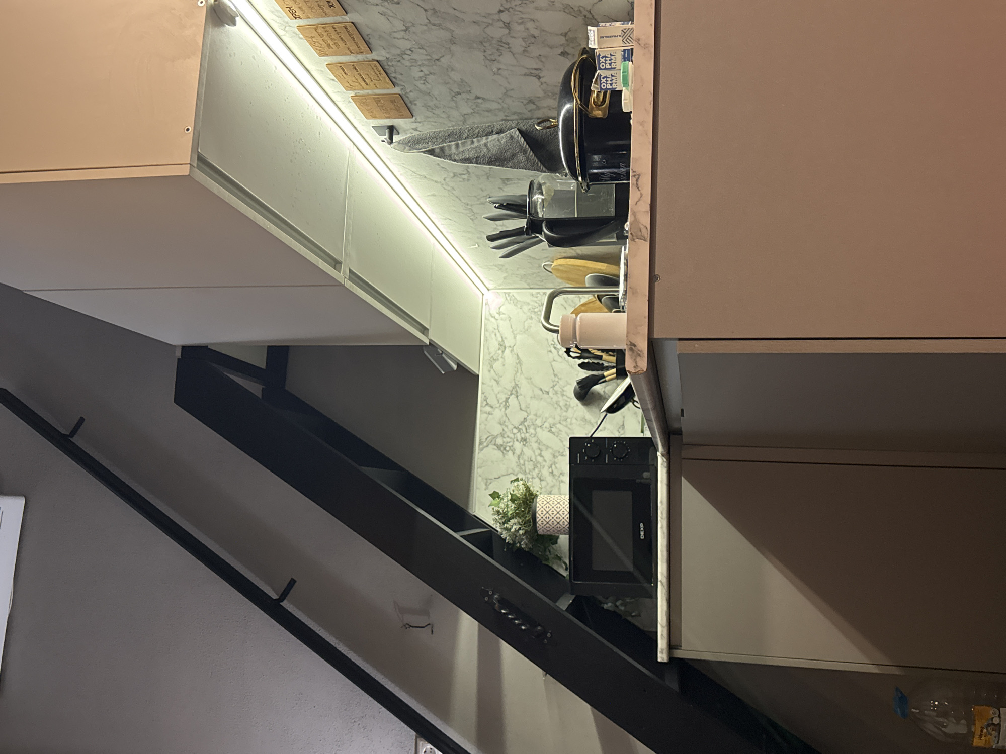 This rotated image displays a contemporary kitchen area, likely indoors during the evening or night given the prominent artificial lighting. No people are visible.
The scene focuses on a kitchen counter with a white and grey marble pattern, extending up the backsplash. Underneath upper cabinets, a bright LED strip light illuminates the counter space. To the right, a light brown/beige cabinet is partially visible.
On the countertop, various kitchen items are arranged. These include a black kettle with gold accents, a clear container holding various utensils (spatulas, whisks), and a white mug. A grey towel hangs near the kettle. Further left, a small decorative plant in a patterned pot is placed next to a black DEXP microwave. On the left side of the frame, a dark-colored structure resembling a staircase or a multi-tiered shelving unit with open compartments is visible, leading downwards and suggesting a multi-level living space.
Notable details include:
* **Lighting:** The LED strip provides strong, direct illumination to the workspace.
* **Aesthetics:** The kitchen features a modern design with clean lines and contrasting materials (marble, light wood-look cabinets, dark metal structure).
* **Labels:** On the marble backsplash, several small, rectangular wooden or cork-like items are attached, possibly as decorative elements or labels. Visible text on some includes "XBEX", "500", "7-25", and "150", which could be related to coffee or measurements. A white box near the kettle shows "OXY" and "PHARM.RU" (partially). The microwave brand is "DEXP". A plastic water bottle is partially visible at the bottom right, with some indistinct text on its label.
The setting suggests a compact, well-organized kitchen space within a home or apartment in Khimki, Russia.