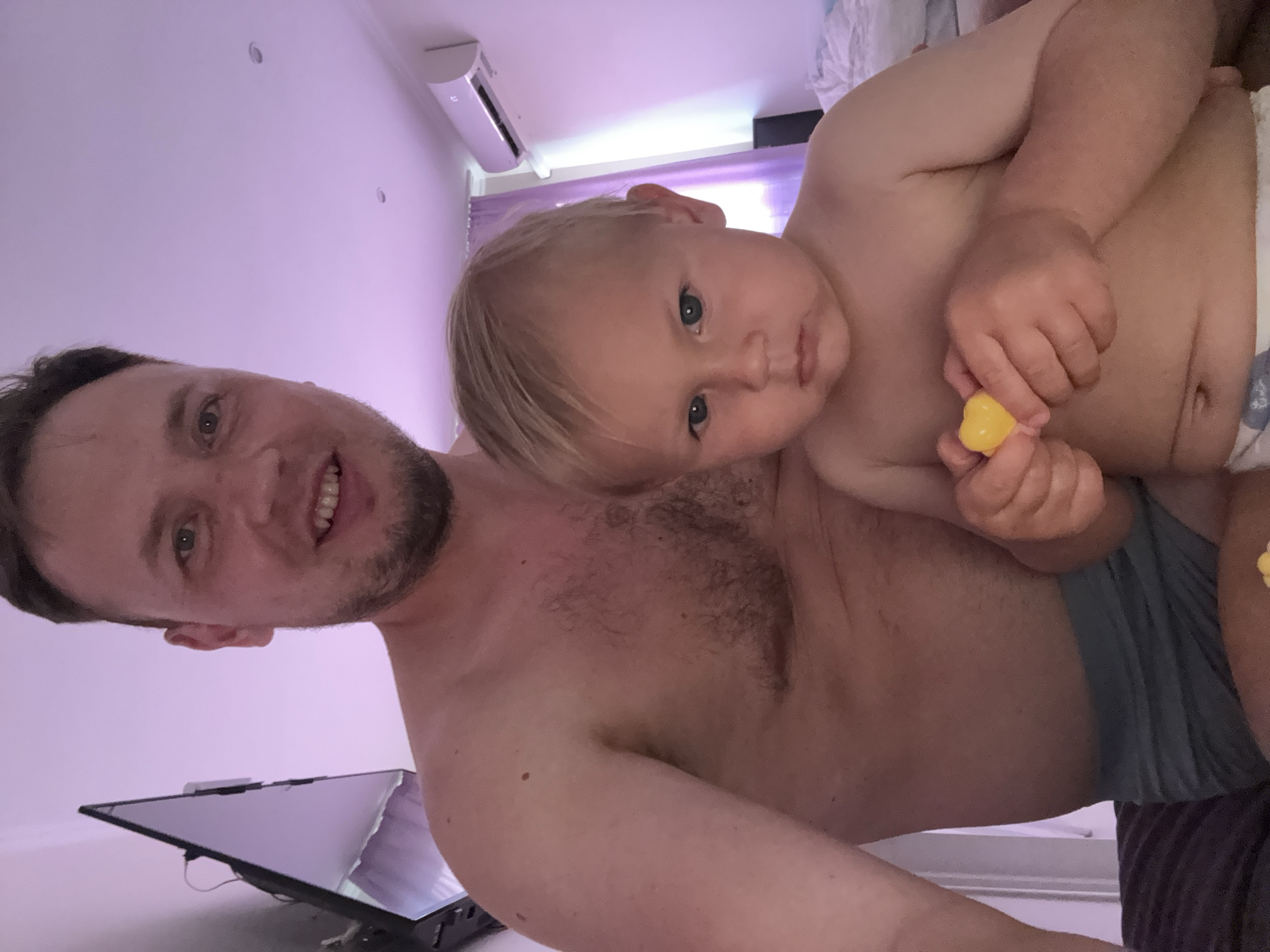 The image shows a man and a baby, possibly father and son, in a room. The man is to the left, smiling and looking at the camera. He has a beard and is shirtless. The baby is on the man's chest, looking towards the camera with a neutral expression. The baby has blonde hair and is holding a small yellow object in its hands. Both subjects appear to be undressed from the waist up, with the baby wearing a diaper.
The setting appears to be a bedroom or living room. In the background, there is an air conditioning unit mounted on the wall, and a window with purple curtains. A laptop or monitor is visible in the lower left corner of the image. The lighting is soft and purple, suggesting it might be evening or the lights are set to a specific mood. There is no visible text in the image. The overall mood seems to be one of affection and warmth between the two individuals. The image is oriented vertically.