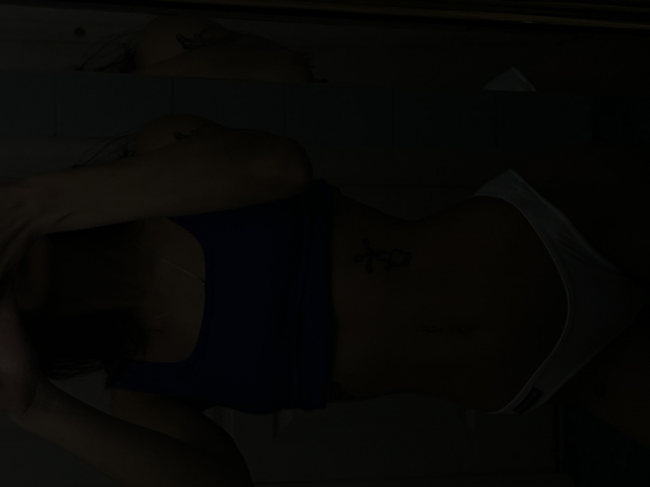 A woman is reclining. She wears a dark blue top and white underwear. Tattoos are visible on her torso. The background is dark, obscuring details.
