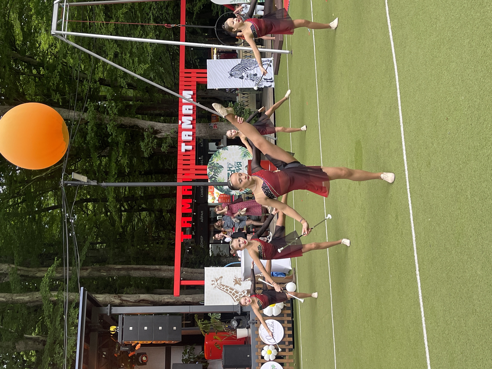 The image depicts an outdoor performance by a group of young girls in matching red and black baton-twirling costumes. They are performing on a bright green turf field marked with white lines, likely a sports or performance area. The girls are in various dynamic poses, with some holding batons.
In the background, there is a stage with sound equipment, red decorative elements with Cyrillic text, and posters featuring animals like giraffes and zebras. A large orange balloon is visible in the upper right portion of the image, and a metal structure, possibly part of an outdoor performance setup or a trapeze, is also present. The scene is set against a backdrop of lush green trees, suggesting an outdoor park or recreational area within a city. The presence of Cyrillic text on the banners indicates a location in Russia. Based on the distinctive red architecture and the overall ambiance, this scene is likely from the "Taman" (ТАМАМ) recreational complex located in Stavropol, Russia, known for its themed zones and outdoor entertainment.