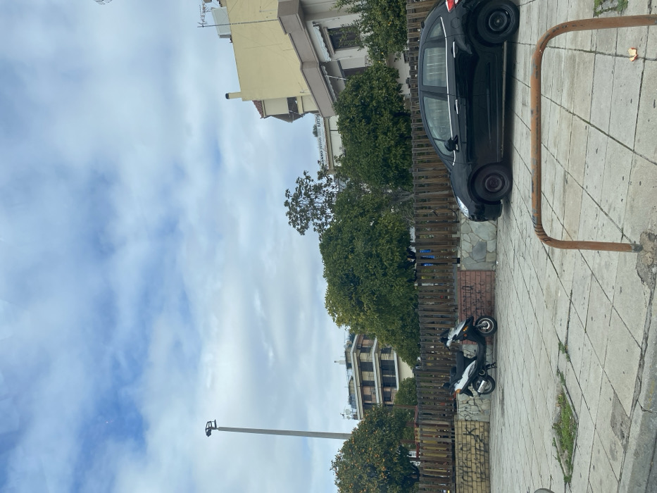 The image captures a quiet, daytime urban street scene in Nikea, Greece. The sky above is largely overcast with scattered lighter clouds.
In the foreground, a paved sidewalk on the right features a rusty, U-shaped metal barrier. A black compact car is parked on an adjacent paved area, partially hidden by a dark wooden fence. Behind this fence, numerous green trees, possibly citrus with small fruits, line the street. Further back and to the left, a black and white scooter is parked on a tiled surface.
Buildings form the backdrop. On the right, a multi-story building with a light yellow facade and brown trim is visible. To its left, a taller, more modern building with multiple balconies and light-colored walls stands. On a building further in the distance, partial Greek text is visible on a sign or banner, reading "ΟΜΟΡΦΟ ΧΩΡΙΟ" (meaning "Beautiful Village").
A tall, slender grey pole, likely a lamppost or utility pole equipped with a small camera or sensor, extends upwards on the left side of the frame against the cloudy sky. No people are visible, suggesting a calm, everyday atmosphere in this residential or mixed-use area.