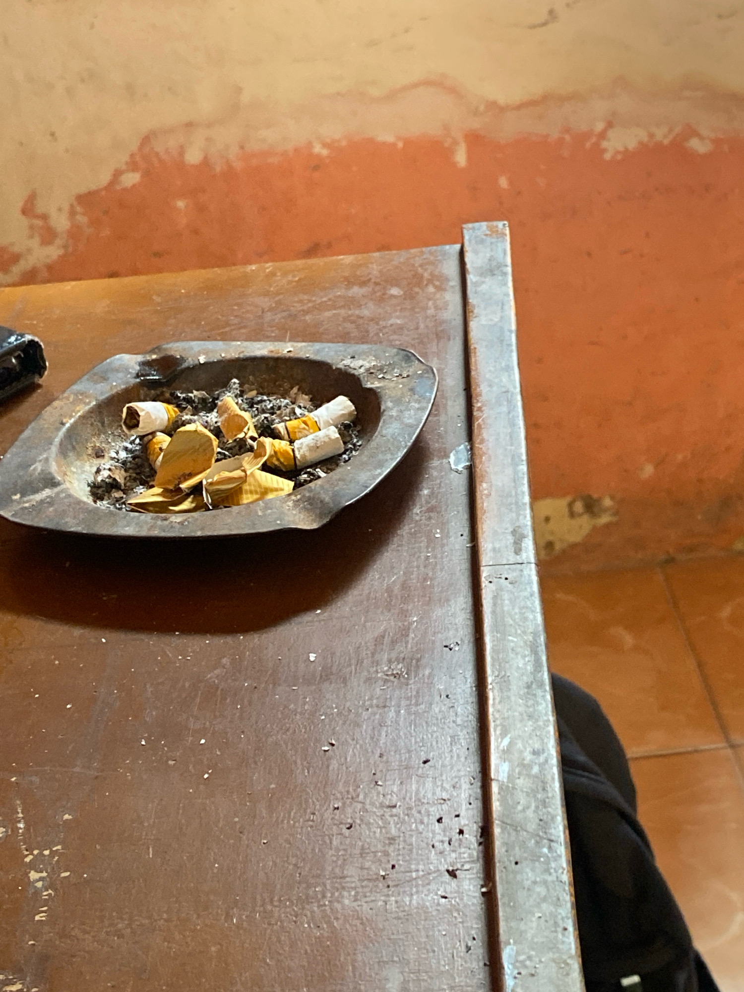 The image depicts a close-up of an ashtray filled with cigarette butts and ash, resting on a wooden table. The ashtray is made of metal and appears old and worn. The table surface is also wooden, showing signs of wear and tear. In the background, a wall with an orange and cream mottled texture is visible. A metal edge of what appears to be furniture or a counter runs vertically along the right side of the image. The scene appears to be indoors, possibly in a casual setting. There is no direct indication or evidence within the image to suggest the location is the city of Mojo Pahit, Indonesia.
